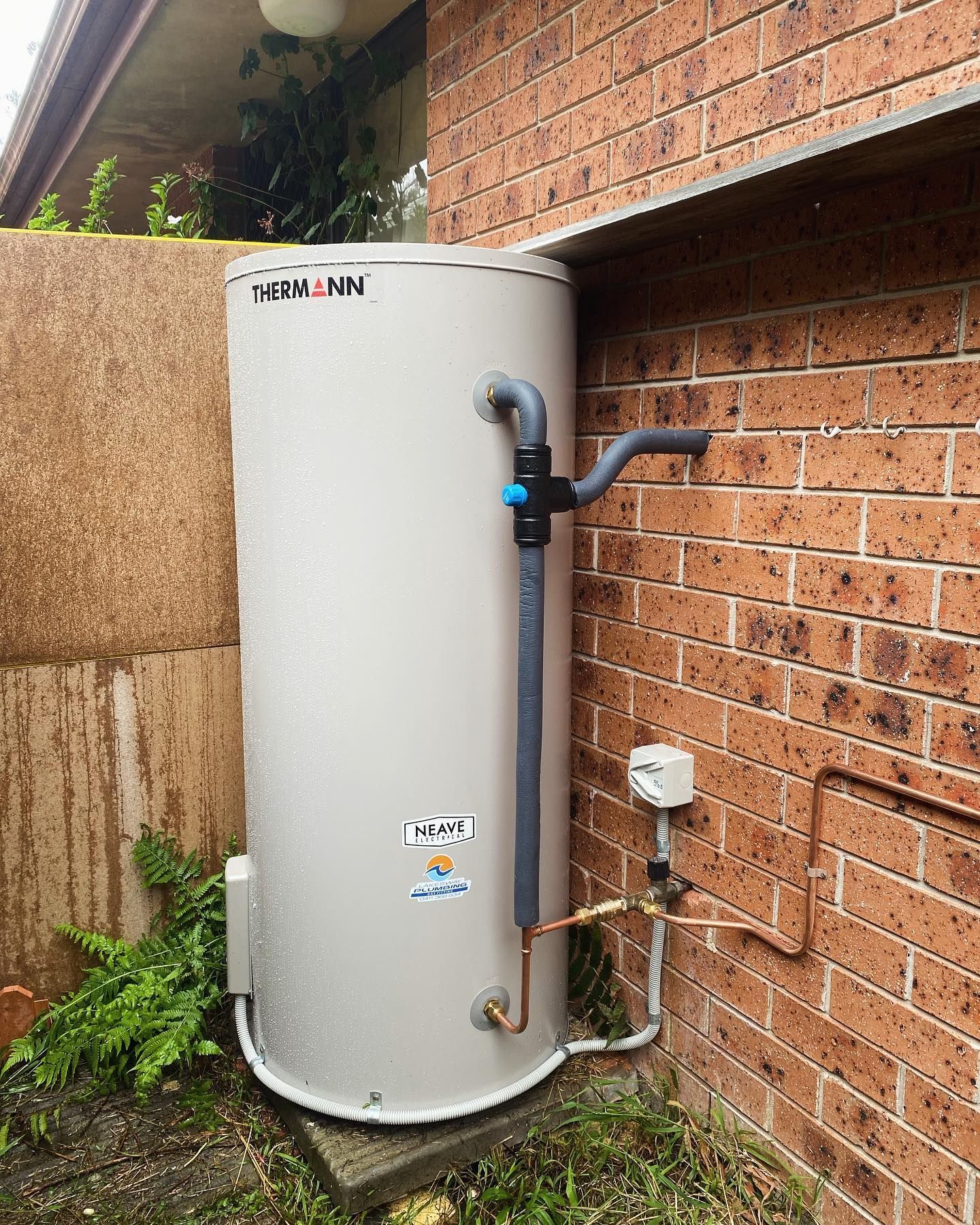 A Cylindrical Hot Water Heater Against a Brick Wall, With Plumbing Visible — Neave Electrical in Forster, NSW