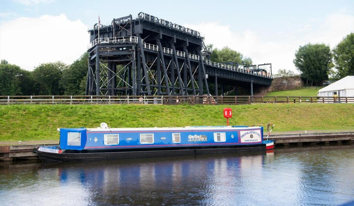Blue canal boat in water near a large black structure, possibly a boat lift, with green grass and trees.