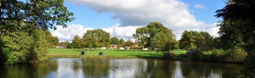 A calm lake reflects a green landscape with trees and a blue sky with clouds.