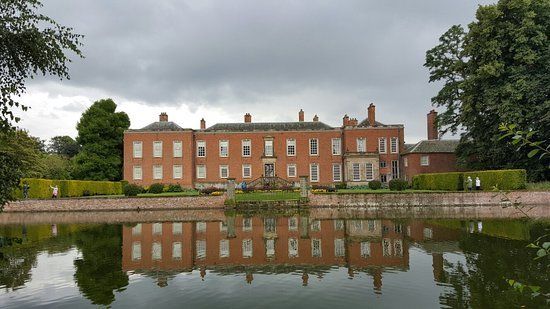 Red brick manor house reflected in calm water under overcast sky.