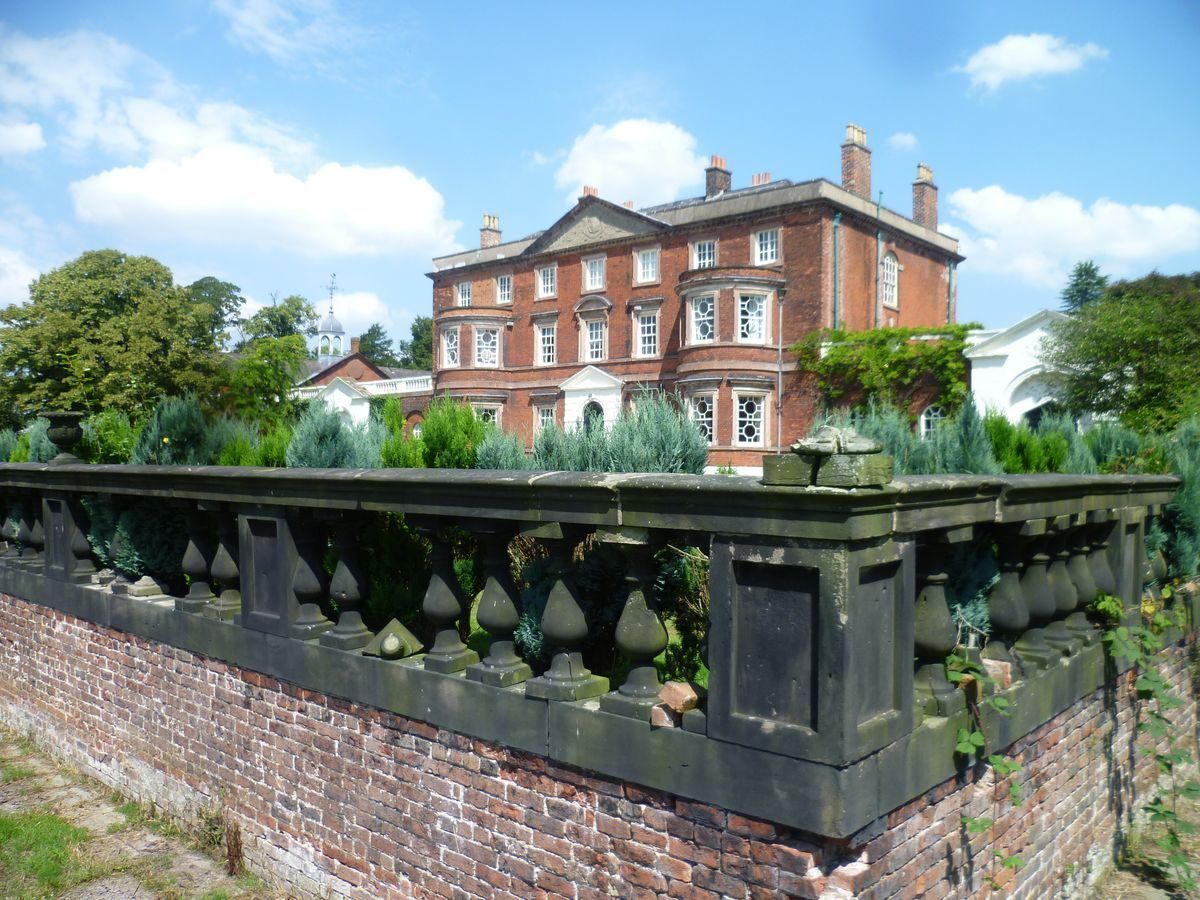 Red brick mansion behind a stone and brick wall, under a blue sky with clouds.