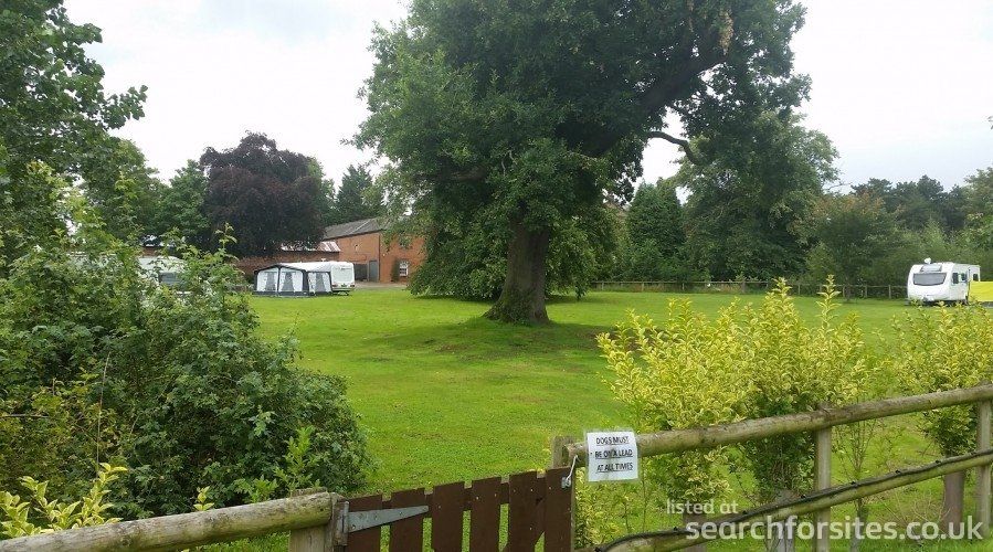 Green grassy campsite with caravans, trees, and a building under a cloudy sky.
