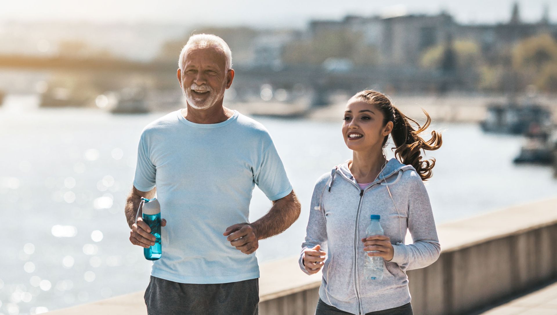 An elderly man and a young woman are jogging by the water.