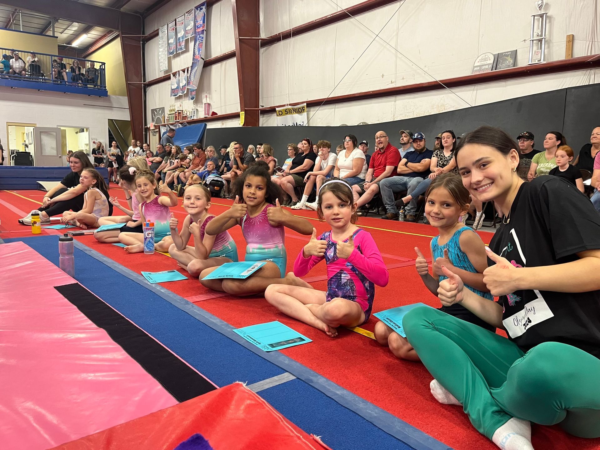 A group of young girls are sitting on the floor in a gym giving thumbs up.