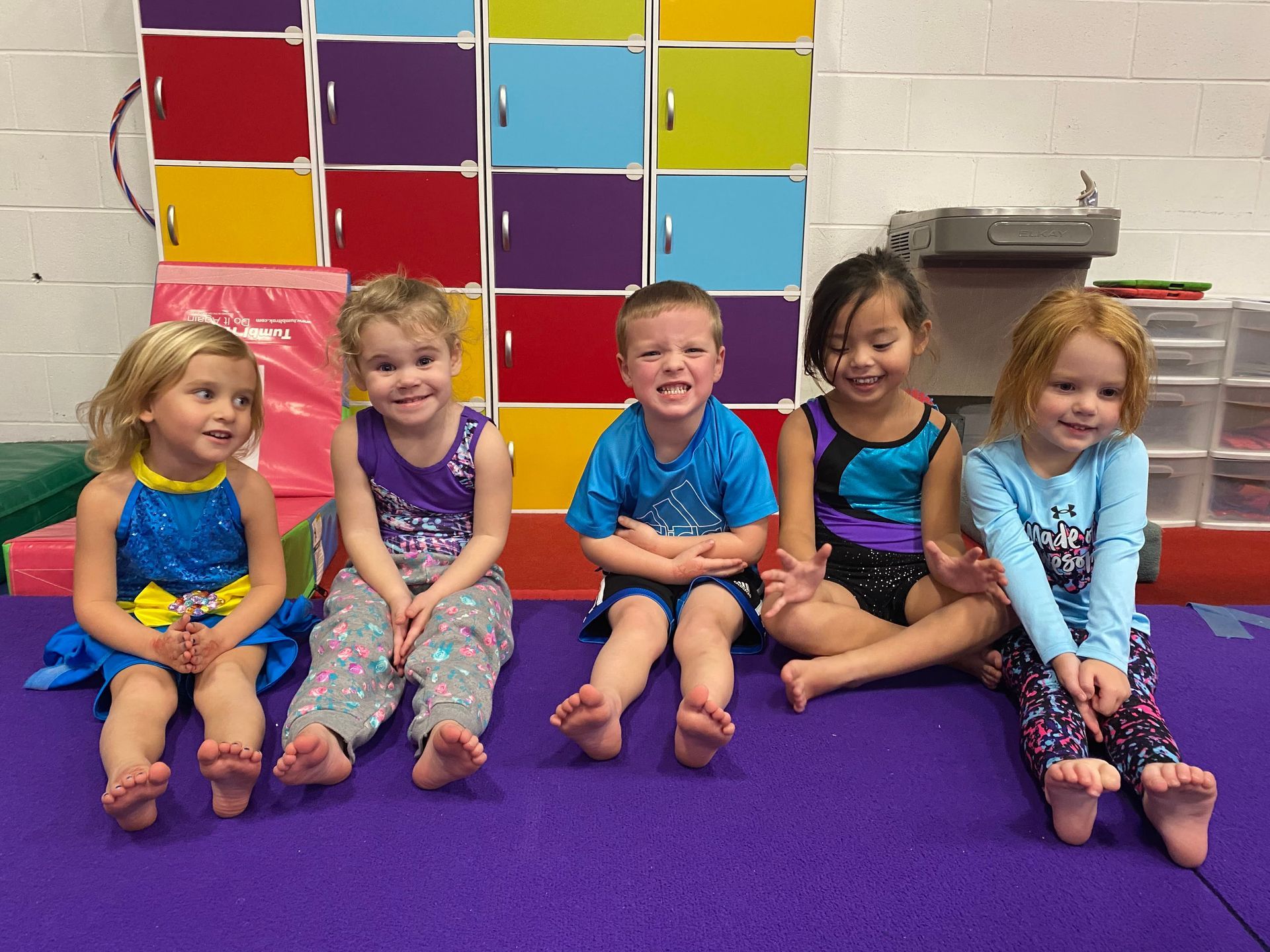 A group of young children are sitting on a purple mat in a gym.