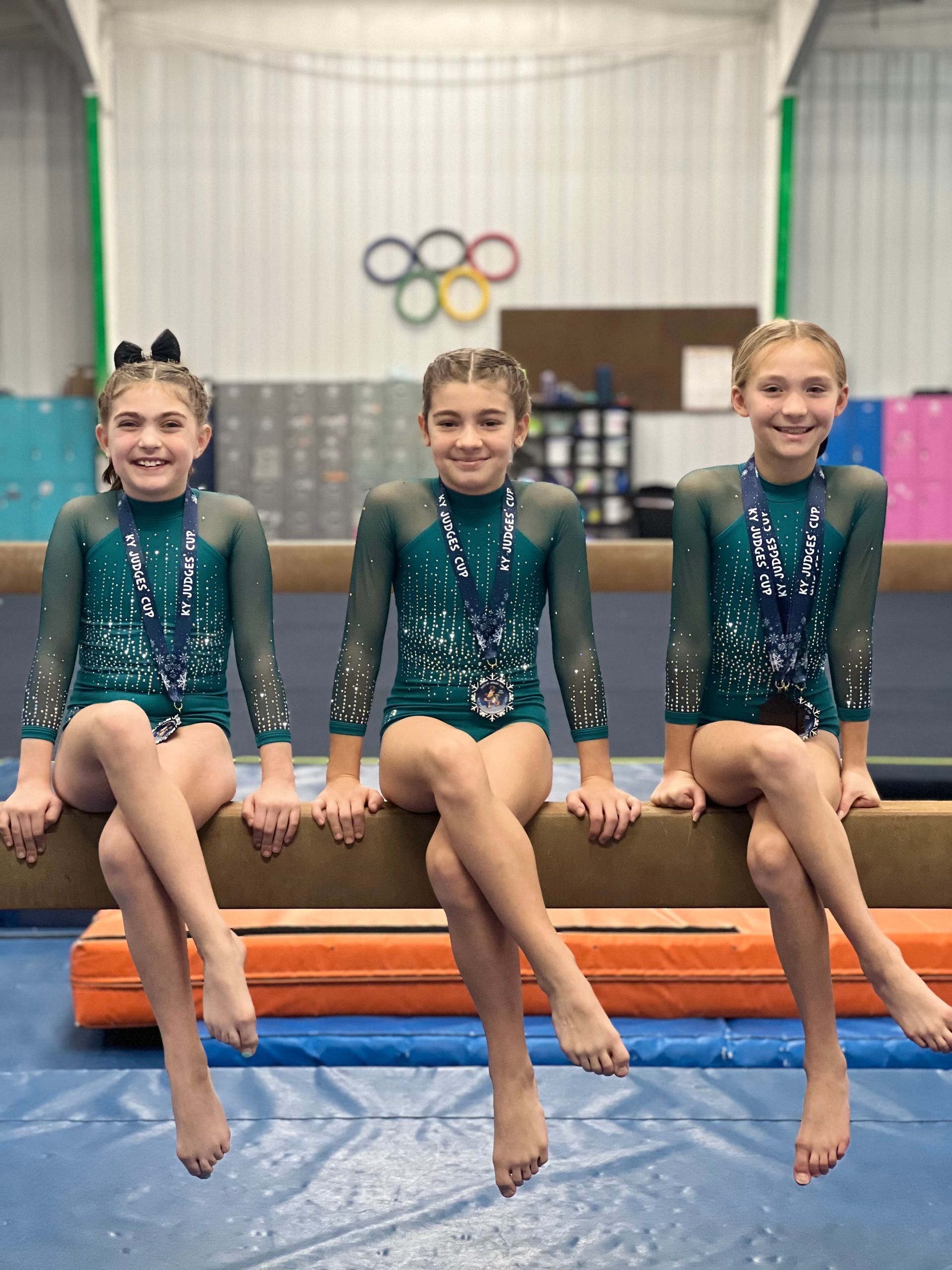 Three young girls are sitting on a balance beam in a gym.