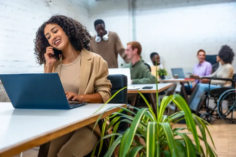 Woman smiling at a laptop in a modern office with plants and people in the background