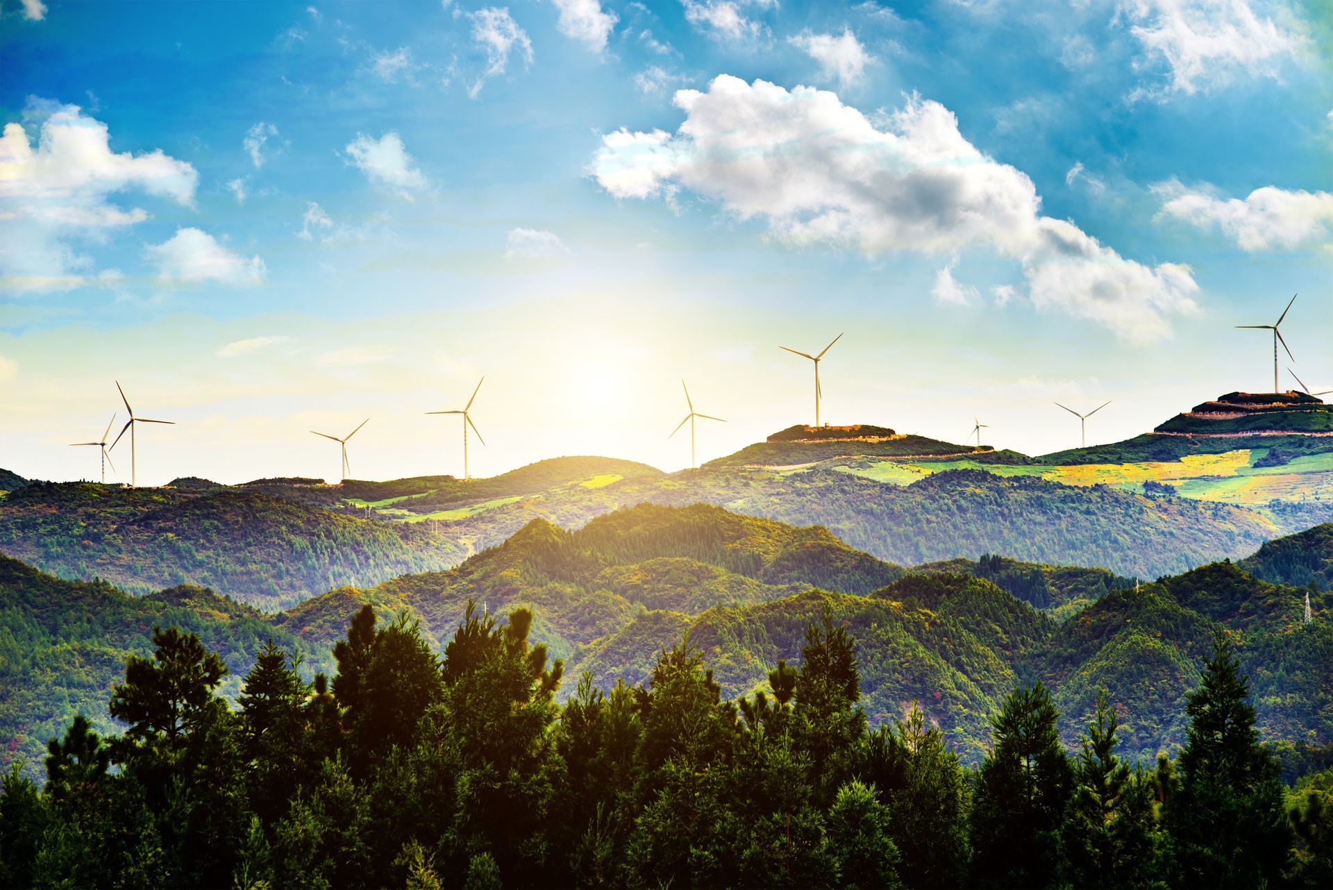 Wind turbines stand atop rolling, tree-covered hills under a bright, sunny sky with scattered clouds.