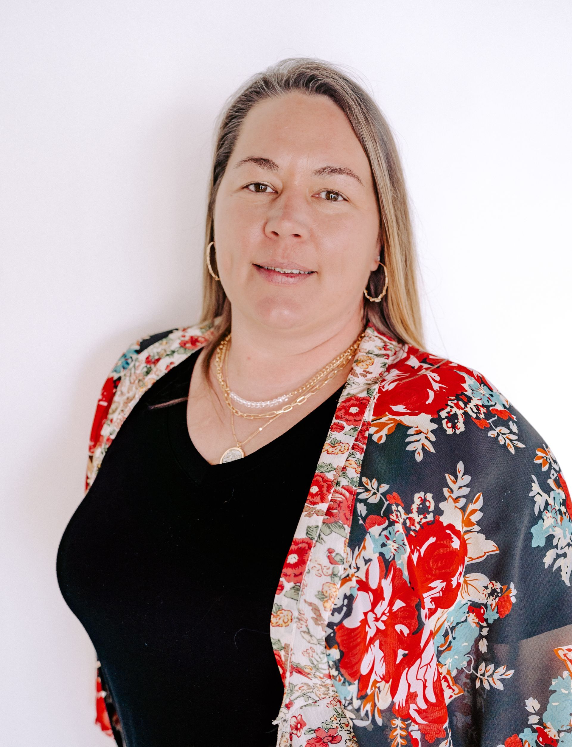 A woman wearing a black shirt and a floral jacket is standing in front of a white wall.