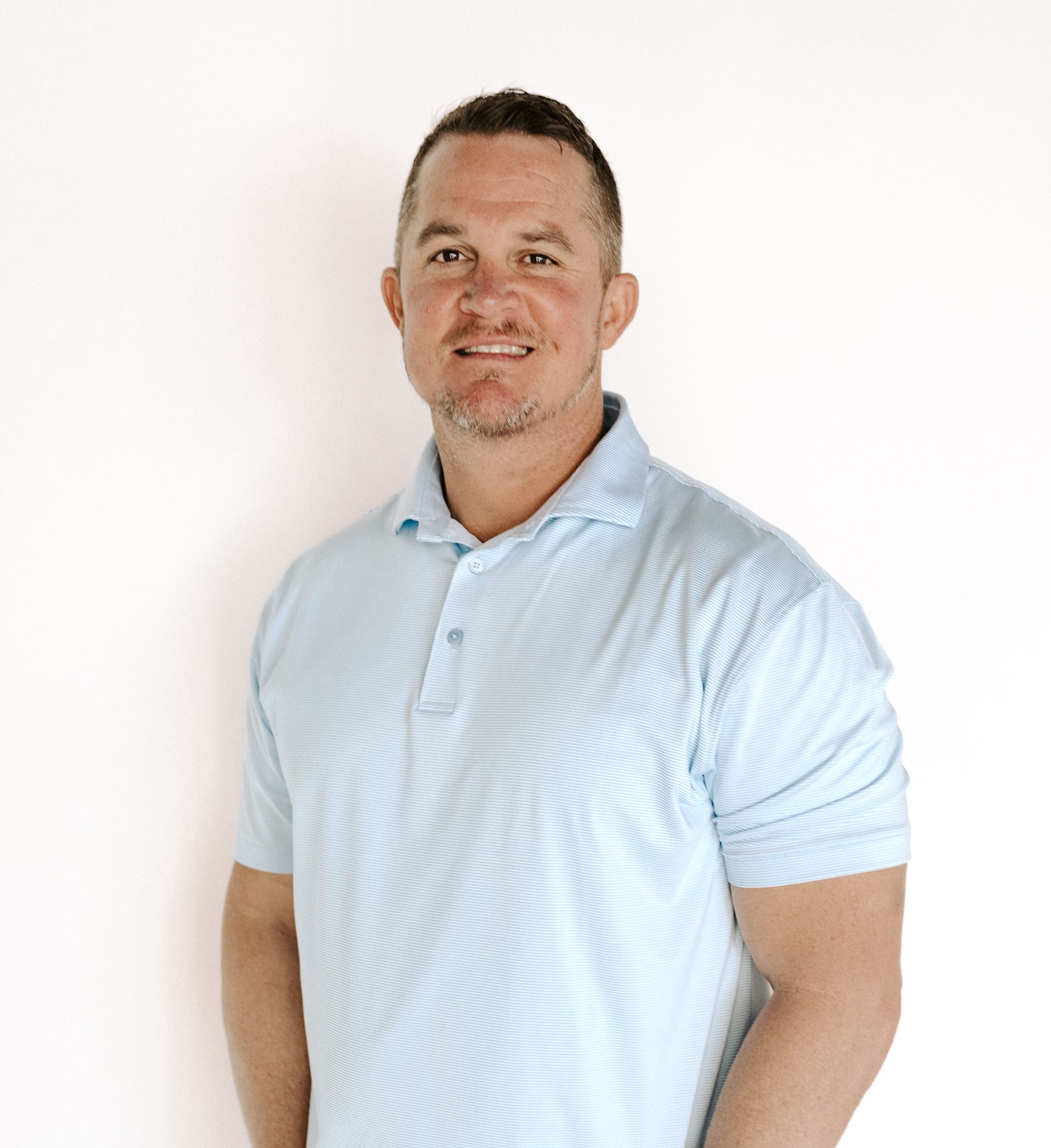 A man wearing a light blue polo shirt is standing in front of a white wall.