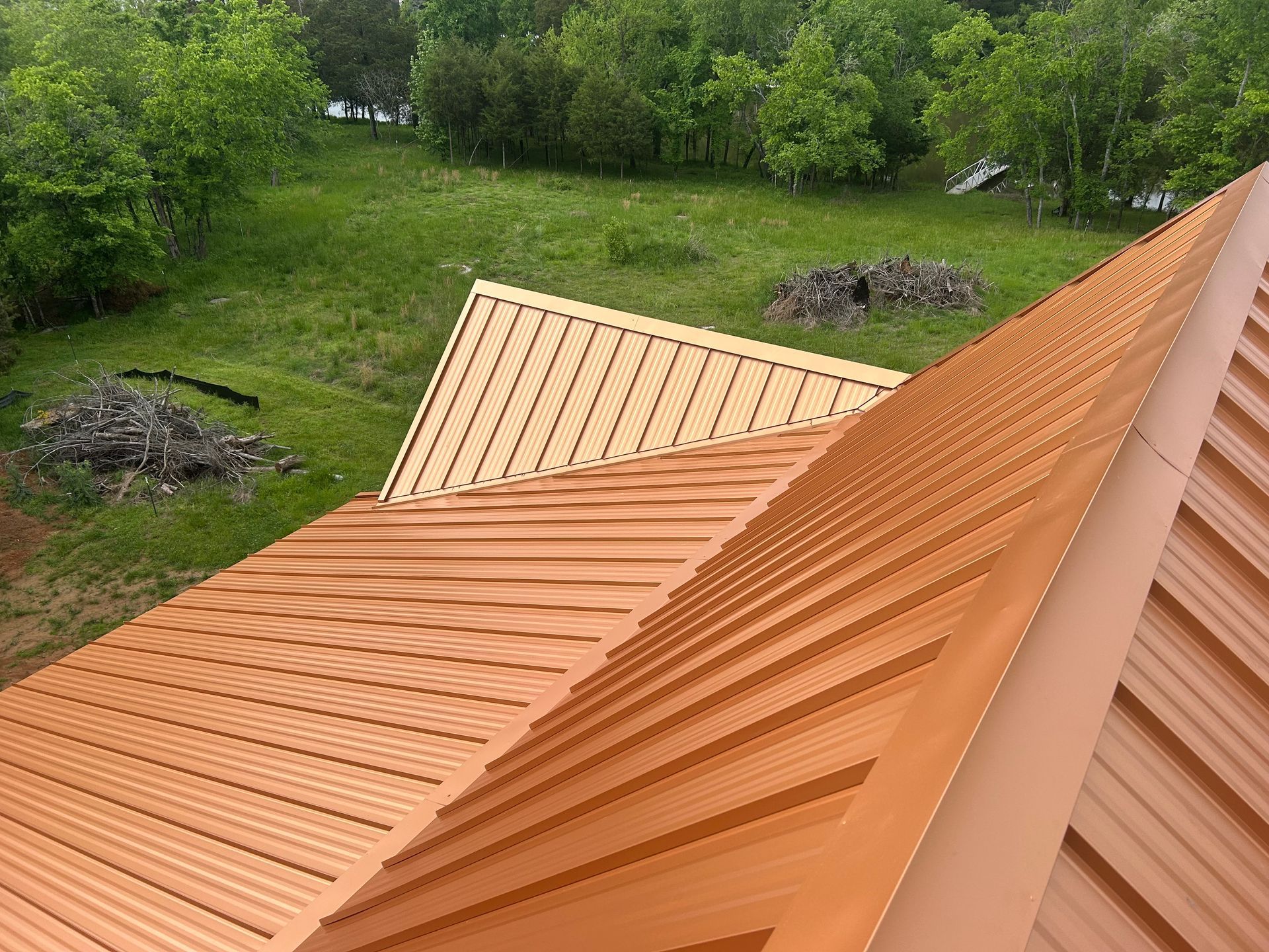 An elevated, angled view of a textured copper-colored metal roof overlooking a lush green yard with trees.