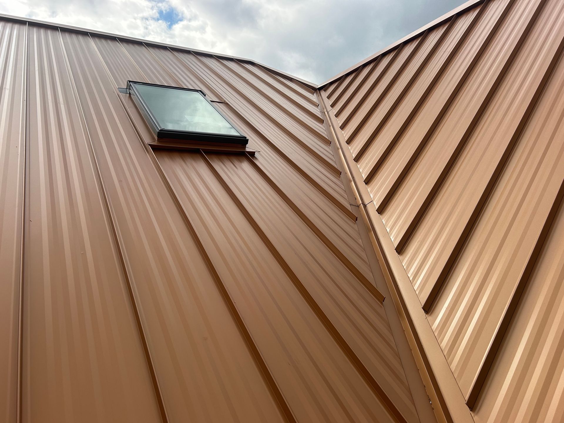A low-angle view of a brown metal roof with vertical seams and a rectangular skylight against a cloudy sky.