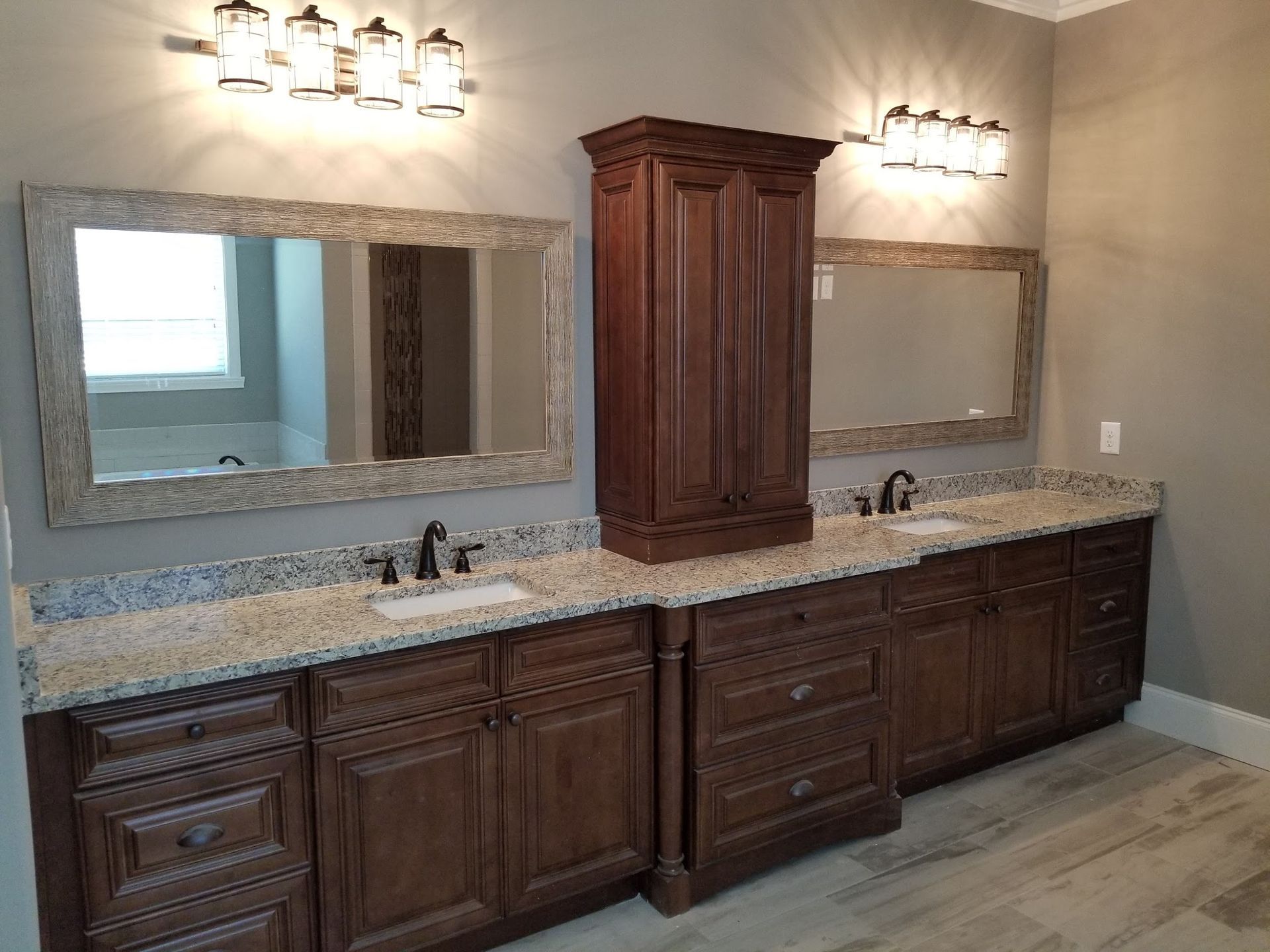 Bathroom with brown cabinets, granite countertops, mirrors, and a tall cabinet. Two sconces above each mirror.