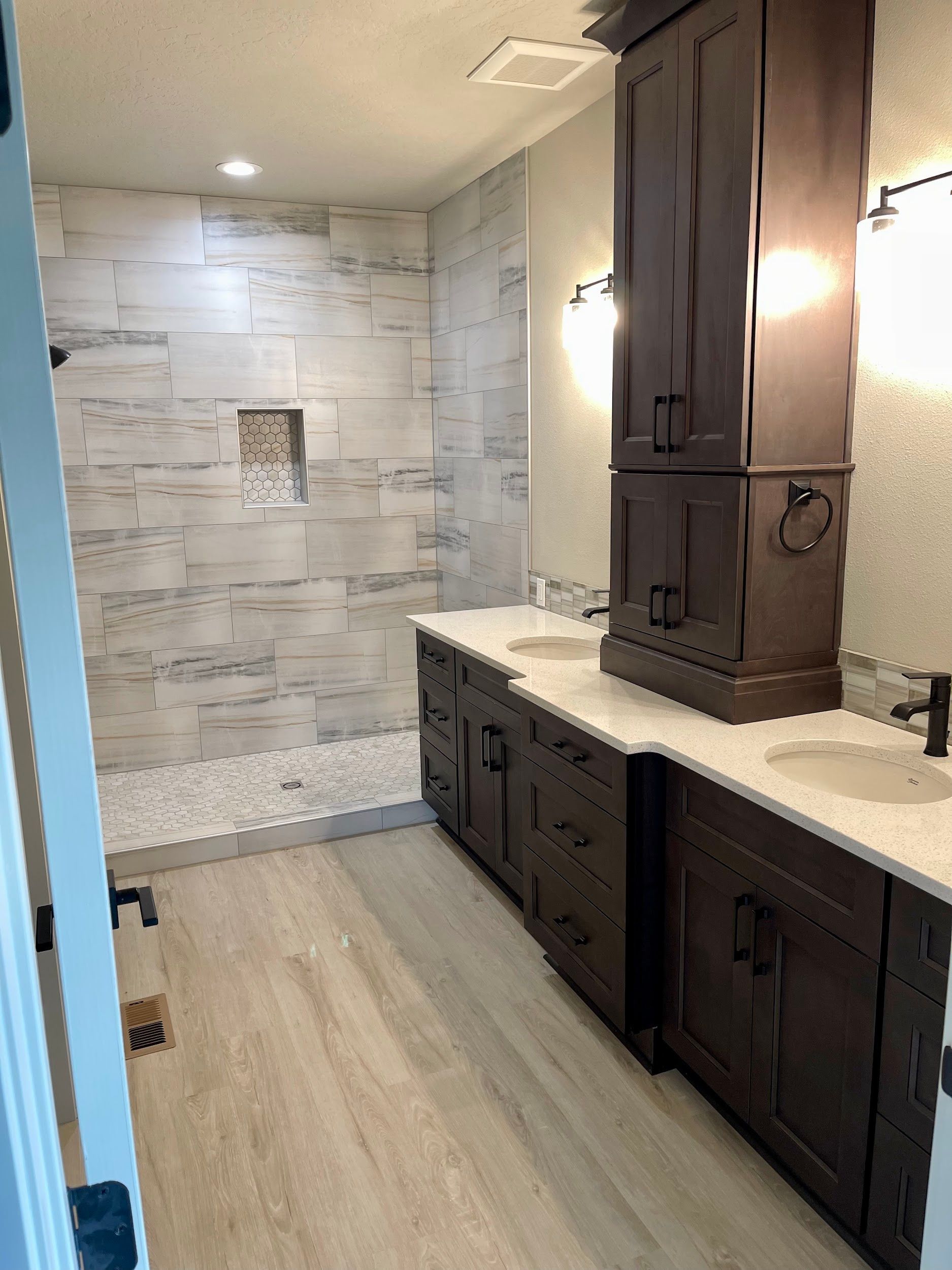 Bathroom with a dark wood vanity, light tile shower, and light wood-look flooring.