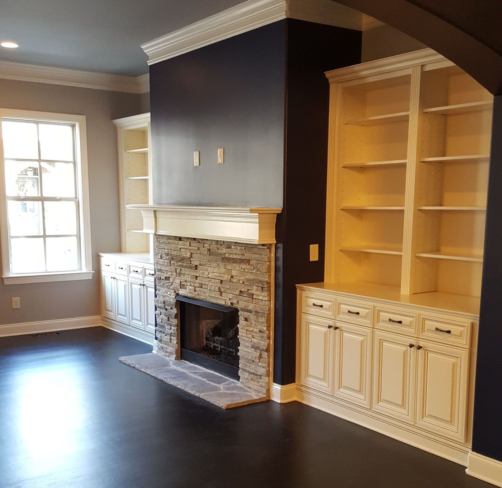 Living room with built-in shelves and stone fireplace, dark walls, white trim and cabinets, dark hardwood floors.