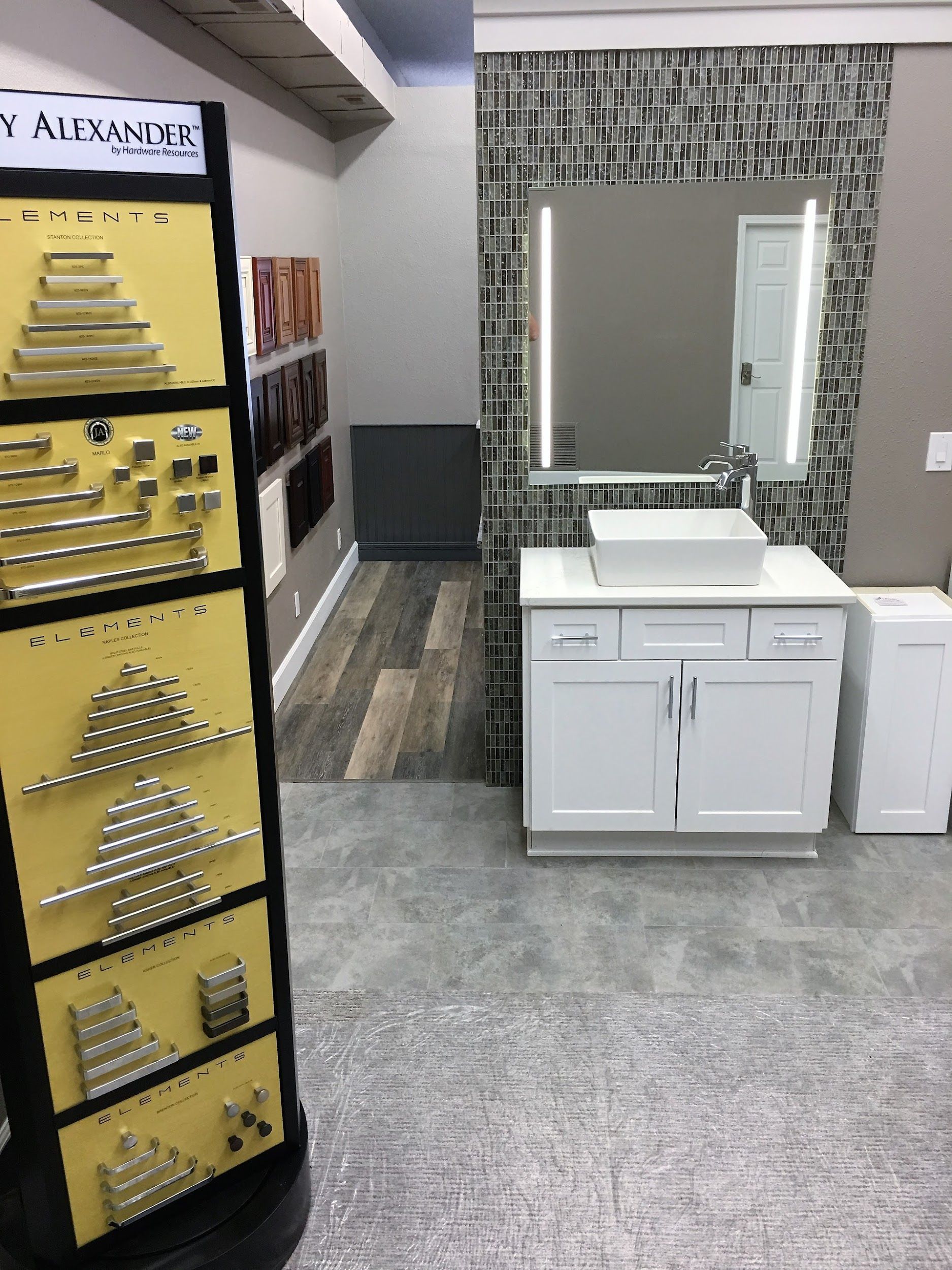 Bathroom showroom with a white vanity, mosaic tile wall, large mirror, and door handle display.