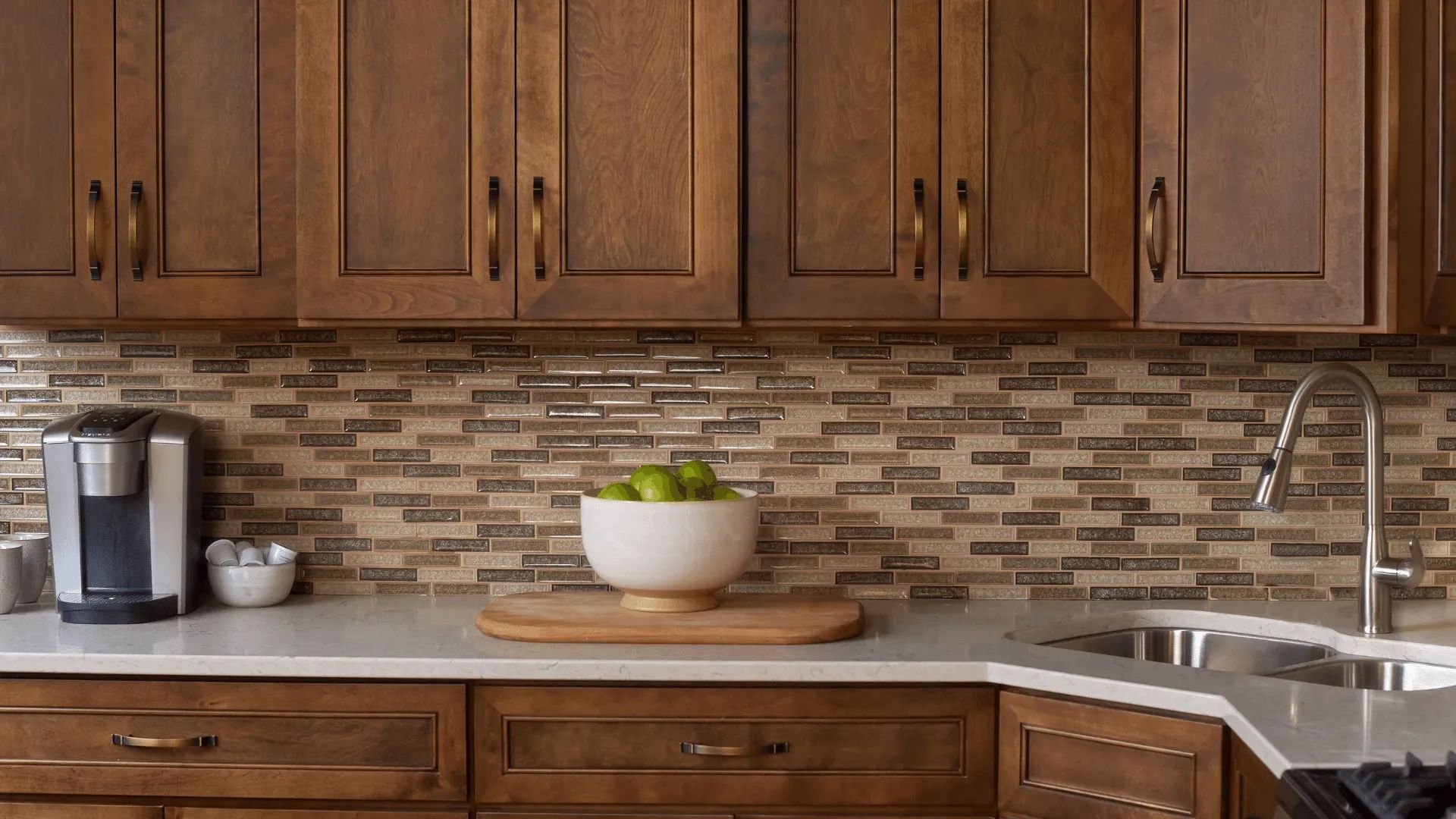 Kitchen with brown cabinets, mosaic backsplash, and countertop with fruit bowl and sink.
