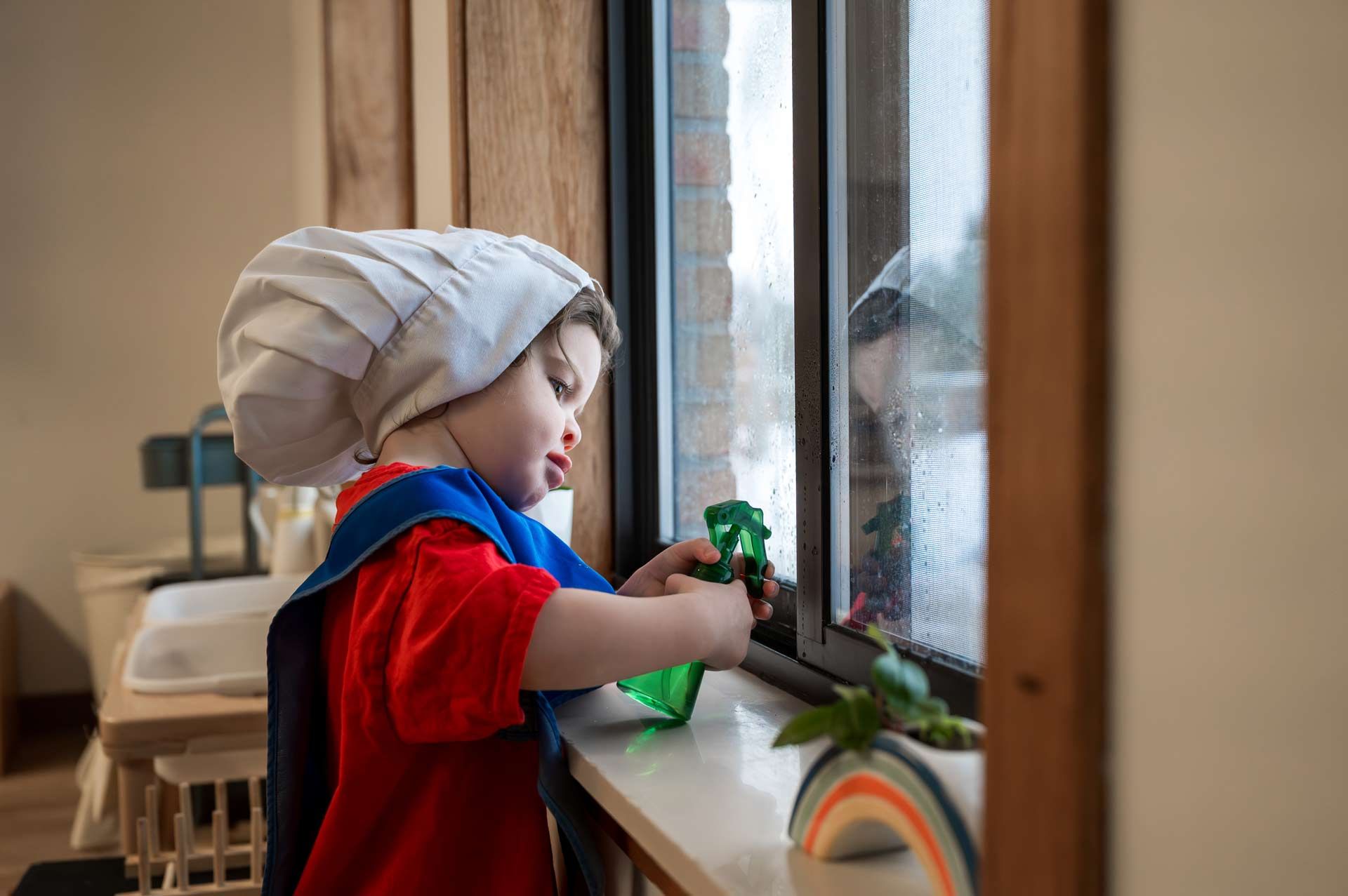 Montessori child wearing a chef hat and using a spray bottle to clean a window