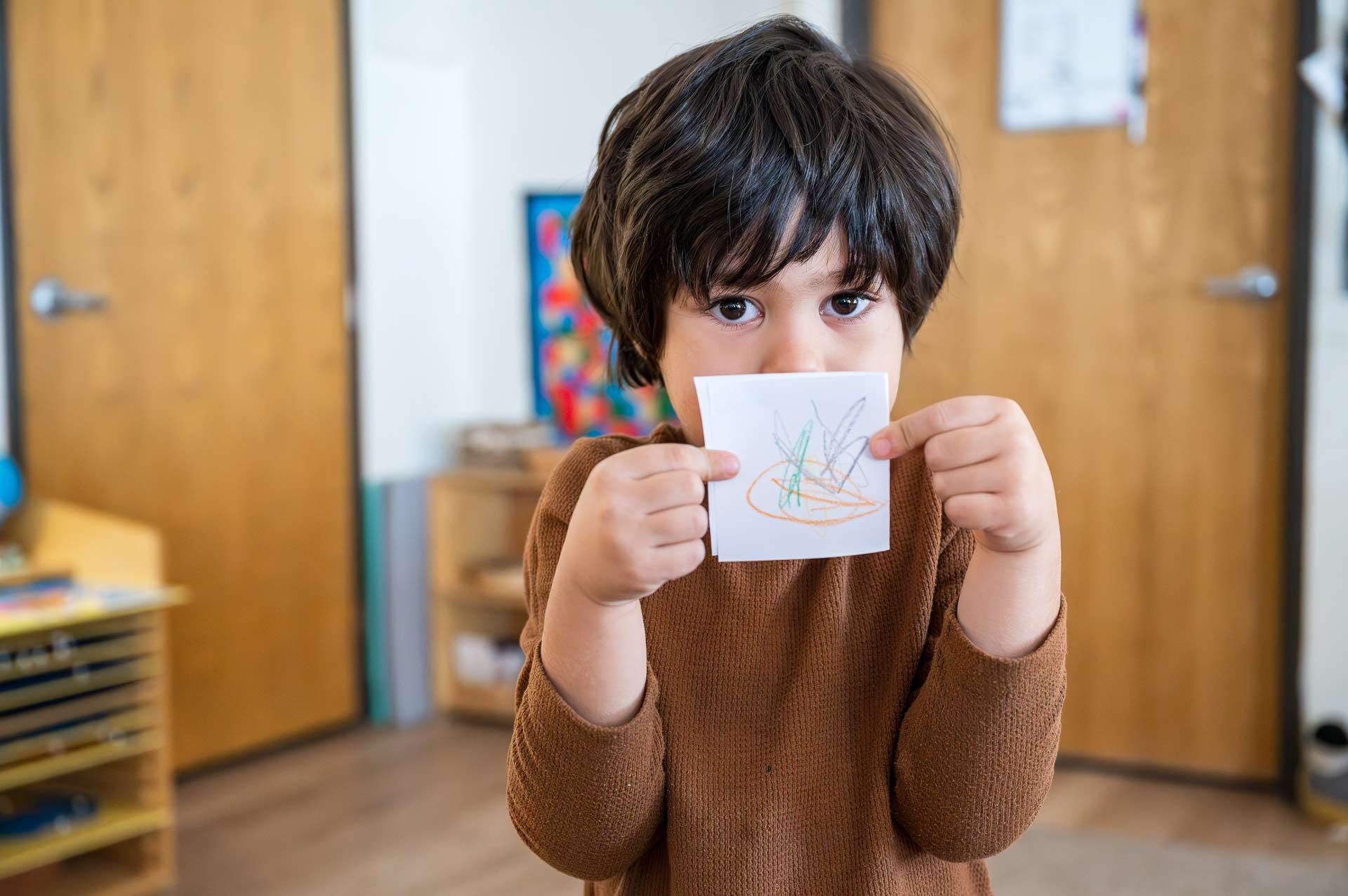 montessori child holding up a crayon drawing in a classroom setting with shelves and artwork in the background