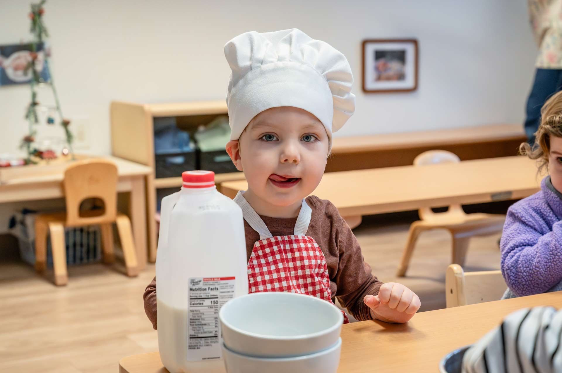 montessori child wearing a chef hat and apron while sitting at a table with a gallon of milk and bowls
