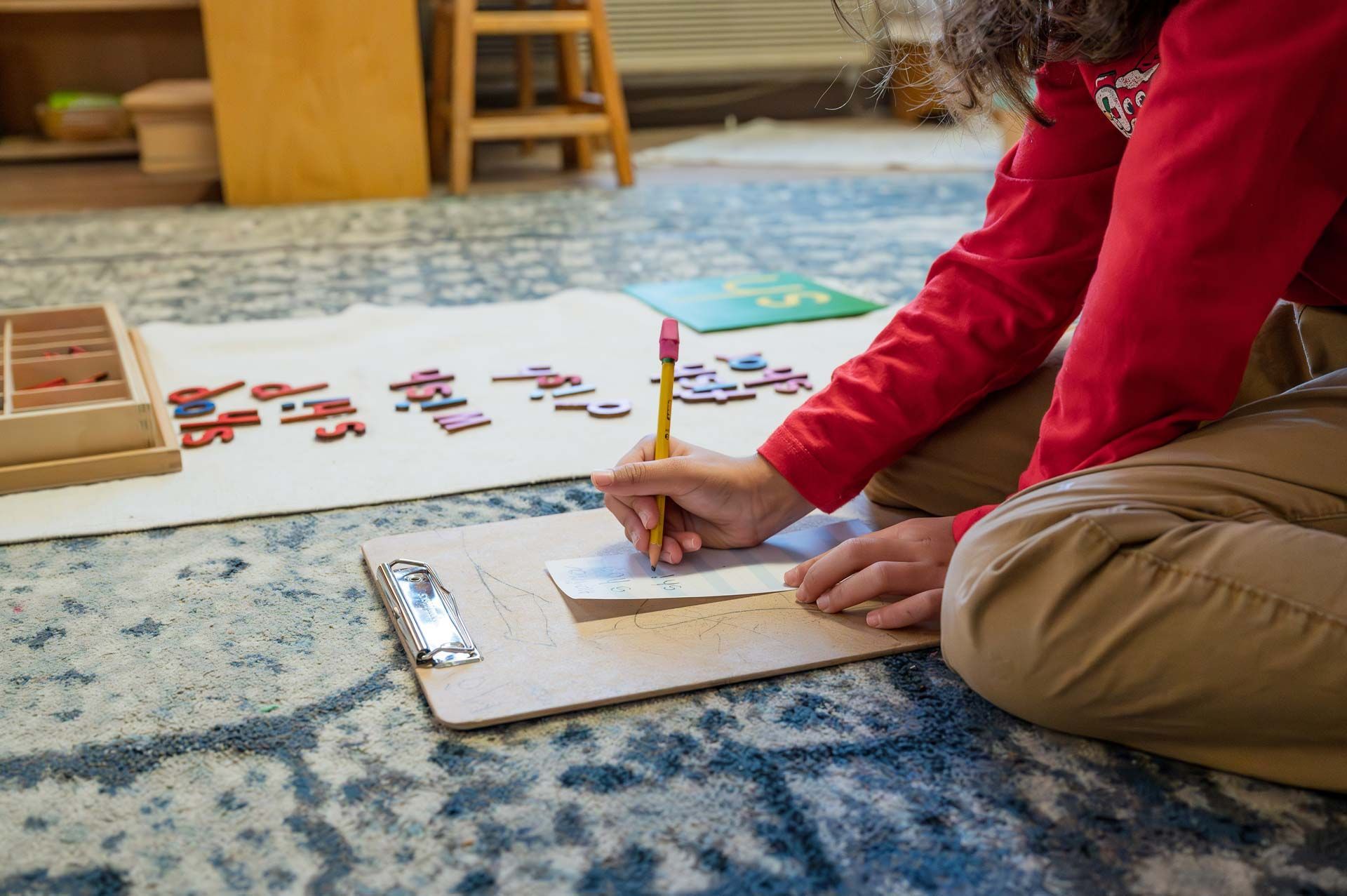 montessori child writing on a clipboard with movable alphabet letters arranged on a mat nearby
