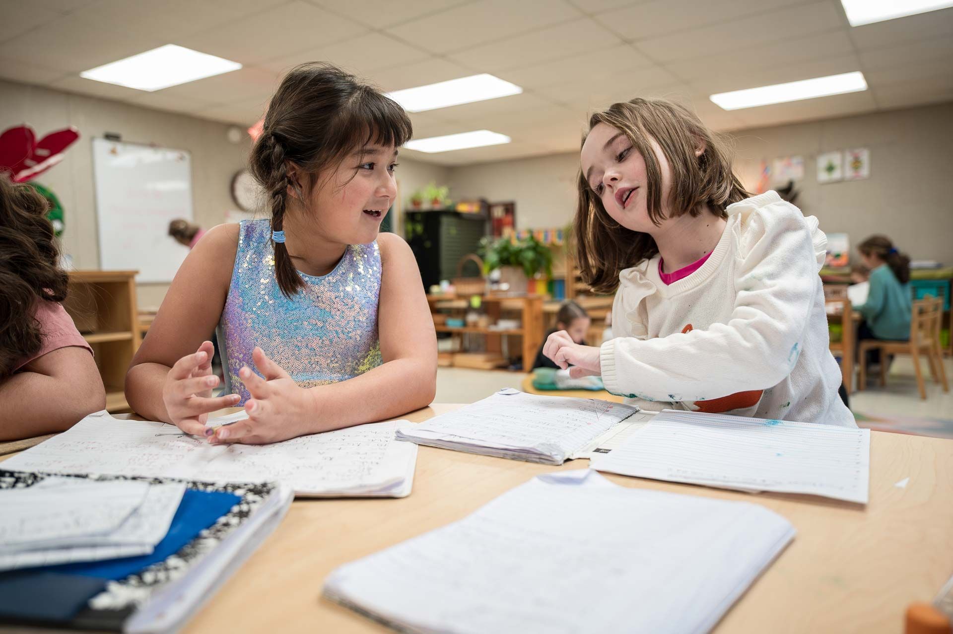 montessori children discussing their work at a classroom table with open notebooks and other students nearby