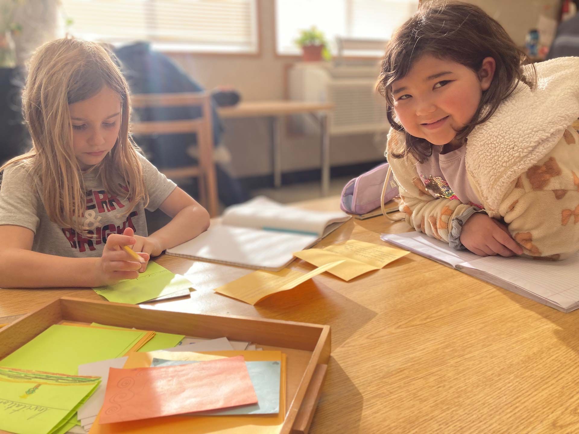two montessori children sitting at wooden table with colored papers and tray natural light from window with potted plant in background