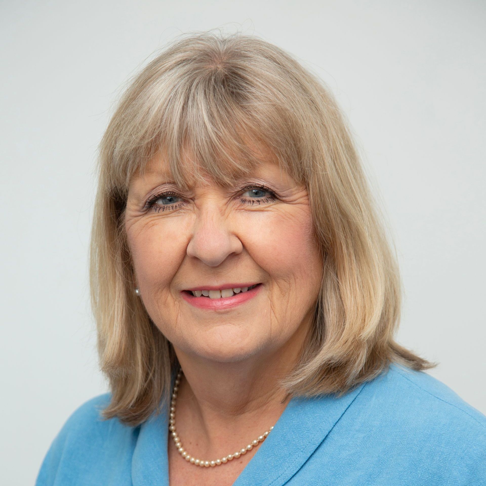 A woman wearing a blue sweater and a pearl necklace is smiling for the camera.