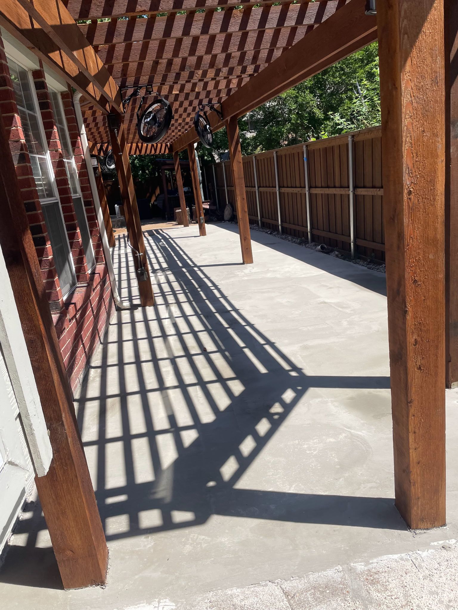 A concrete patio covered by a wooden pergola with shadow lines, next to a brick house wall and a wooden privacy fence.