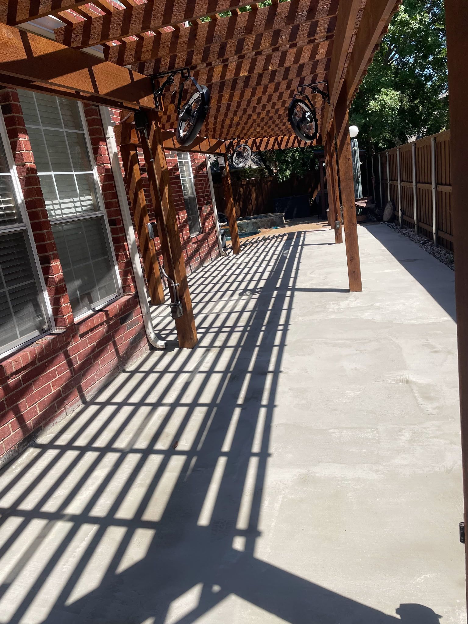 A concrete patio beneath a wooden pergola attached to a brick house, casting long, striped shadows across the ground.