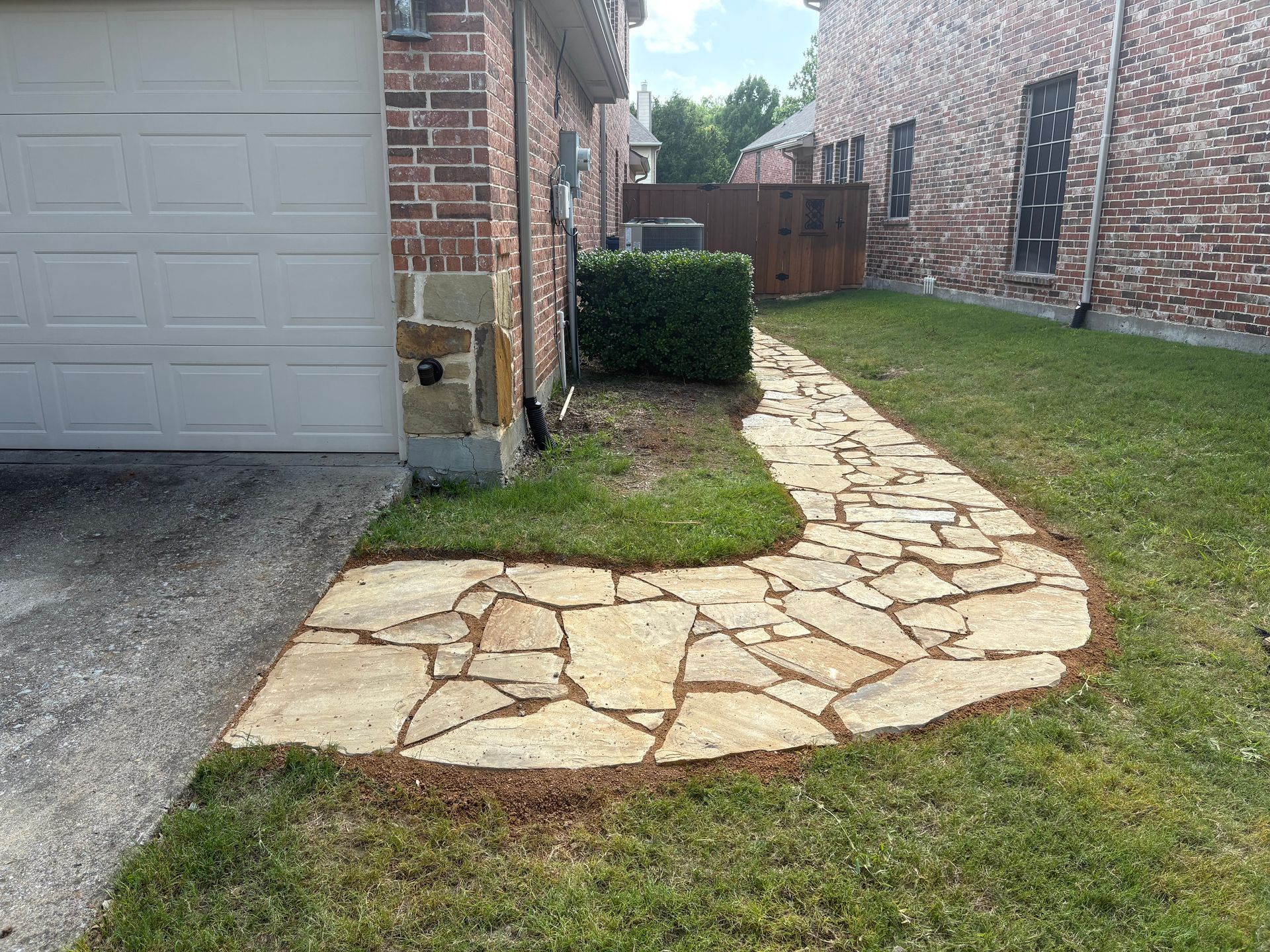 A flagstone walkway curves alongside a house's brick wall and garage, transitioning from pavement onto a grassy lawn.