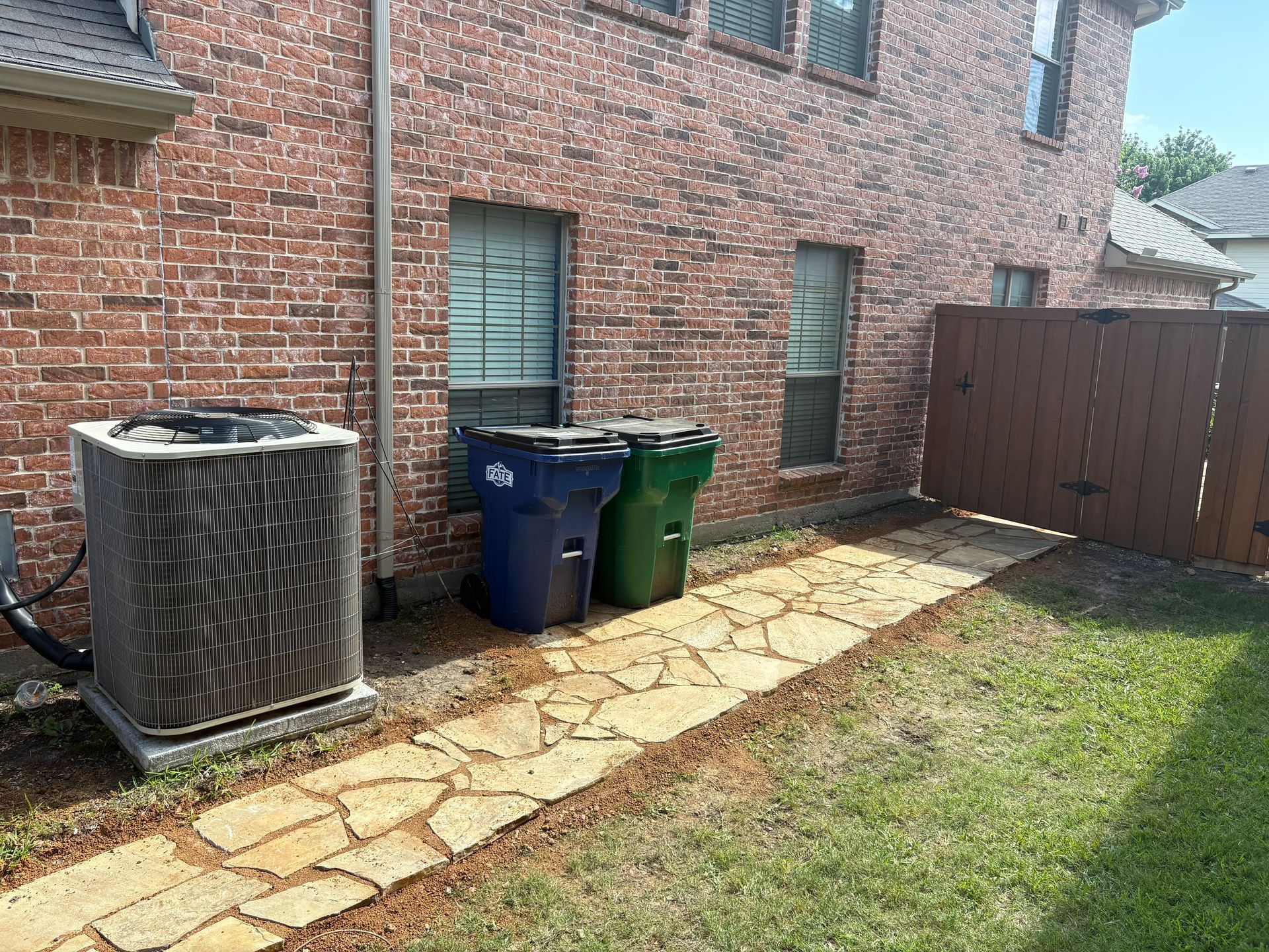 A stone path runs along a brick house wall next to an air conditioning unit and two large trash bins by a wooden fence.
