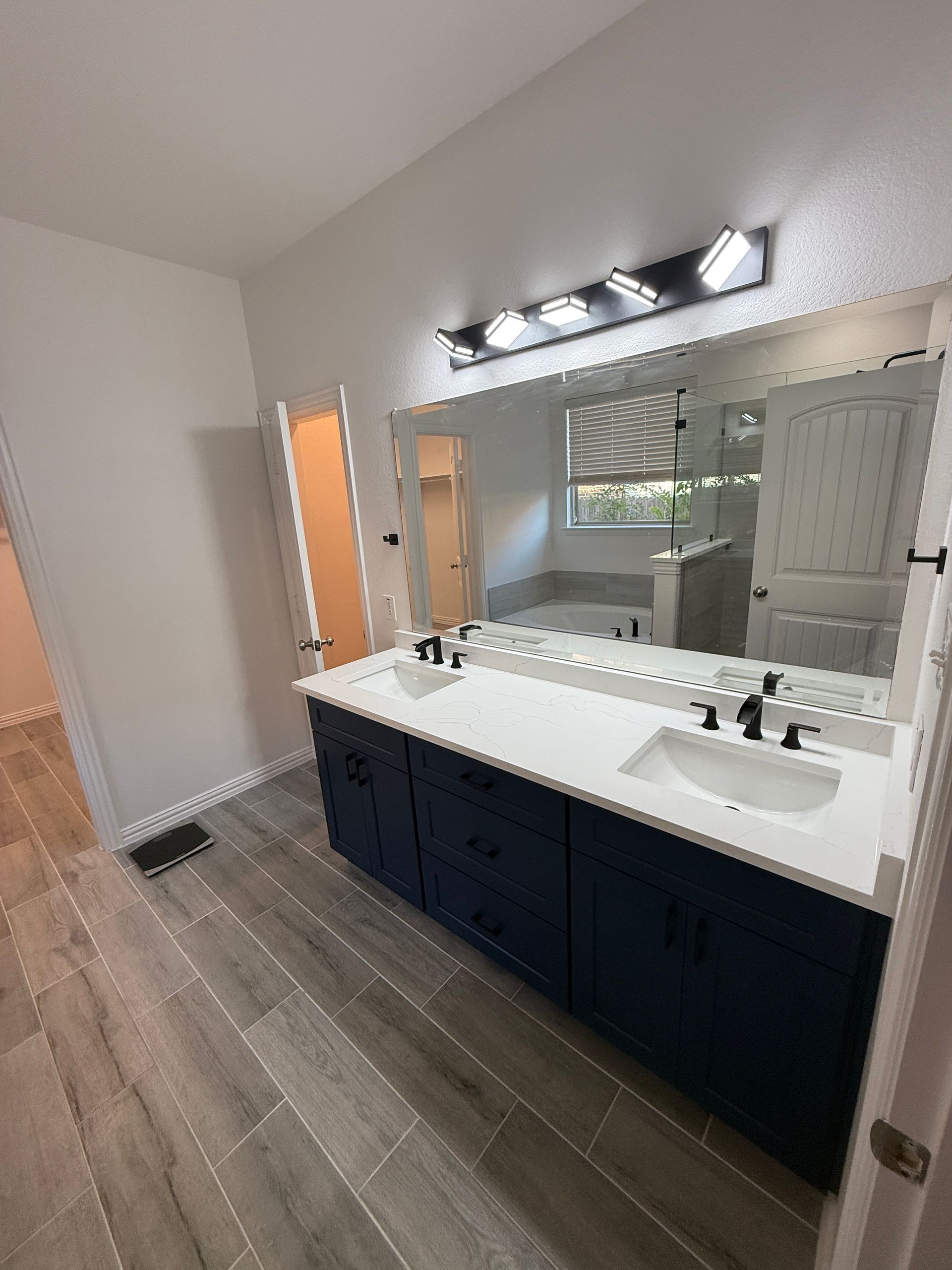 A modern bathroom with a double-sink navy vanity, white quartz countertop, large mirror, and gray wood-look tile flooring.