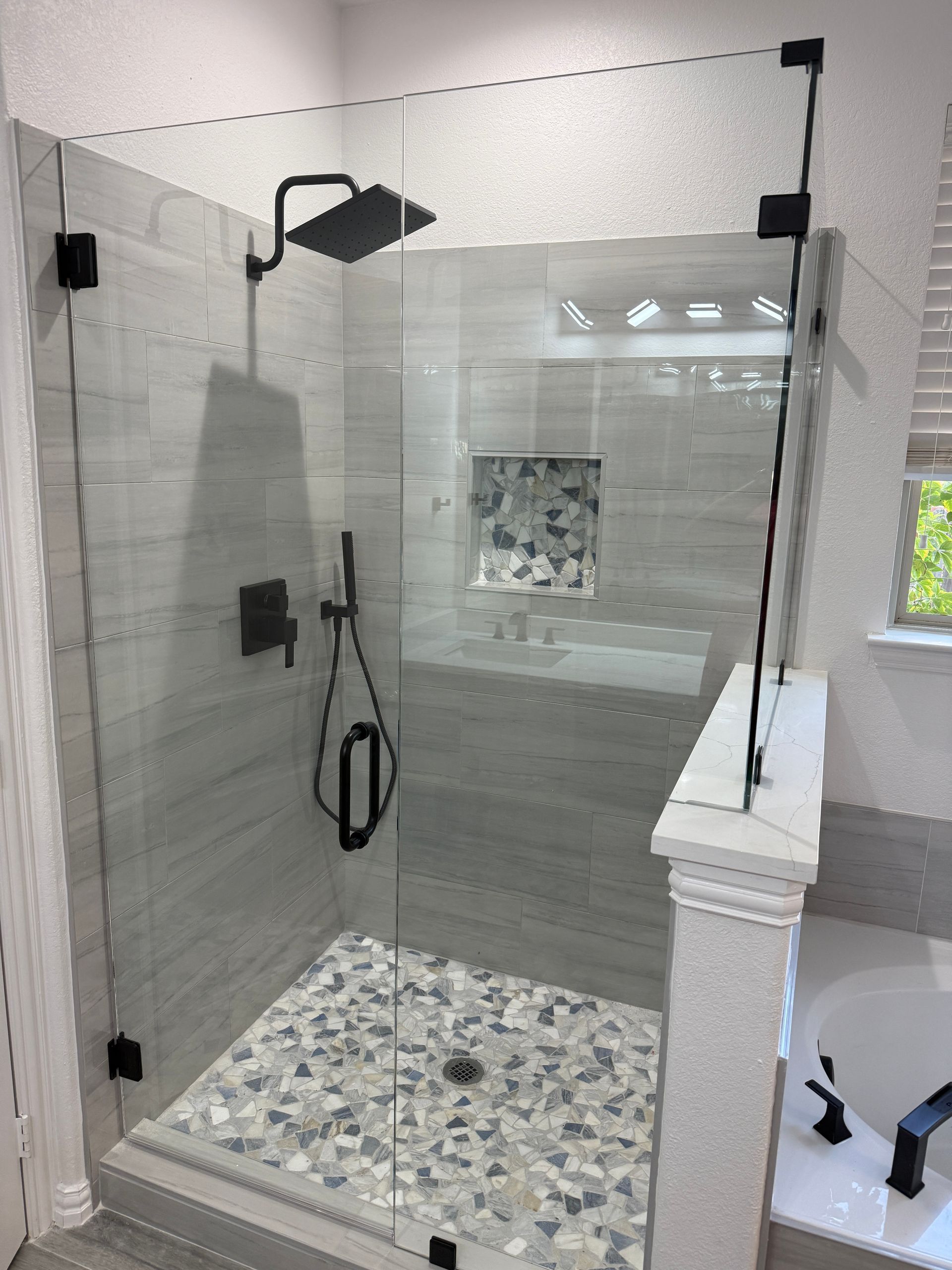 Modern glass-enclosed shower with matte black fixtures, gray tiled walls, and a decorative pebble-style floor.