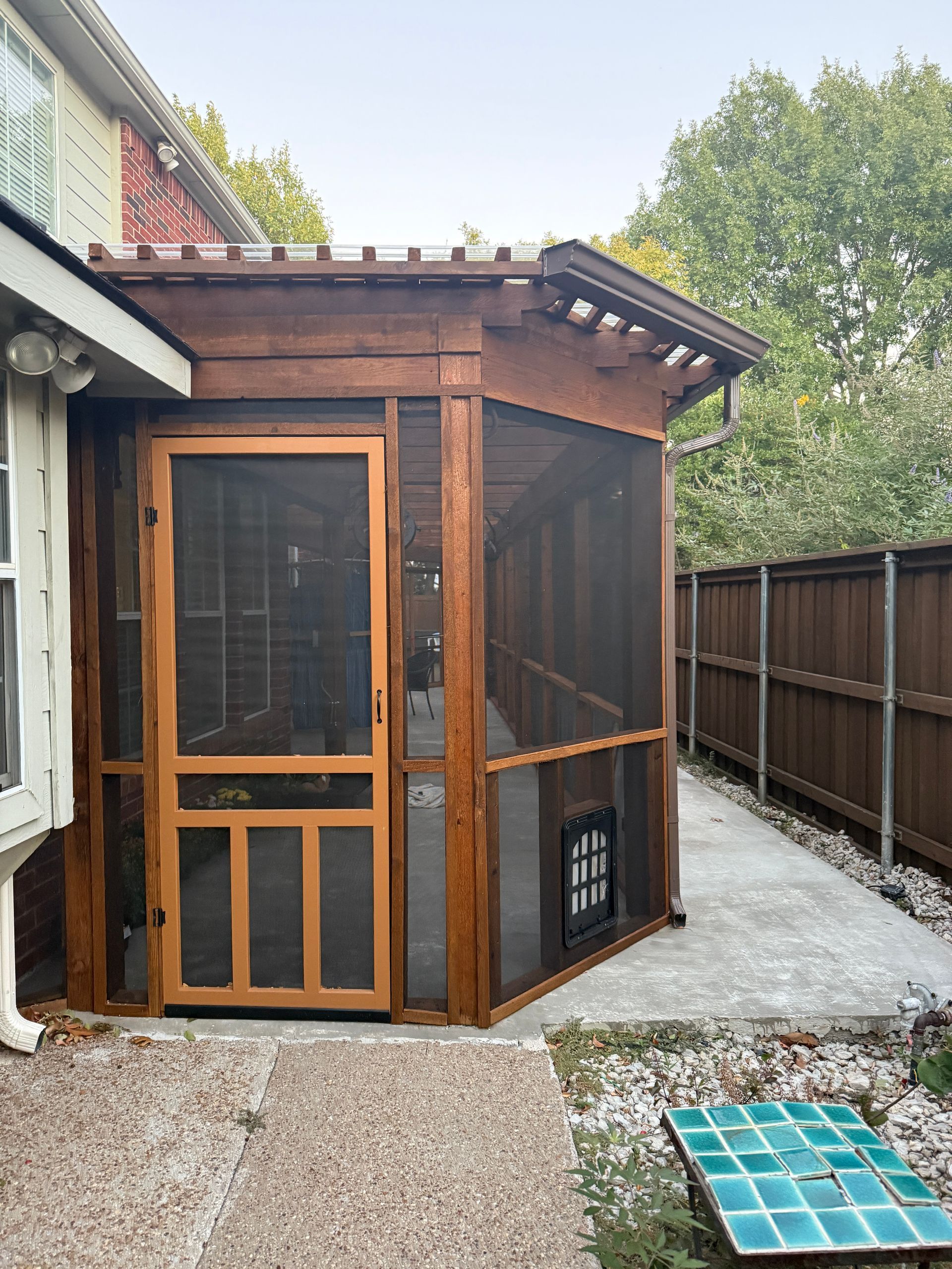 A wooden screened-in porch with a door and a pet door, attached to a house next to a fence in a backyard.