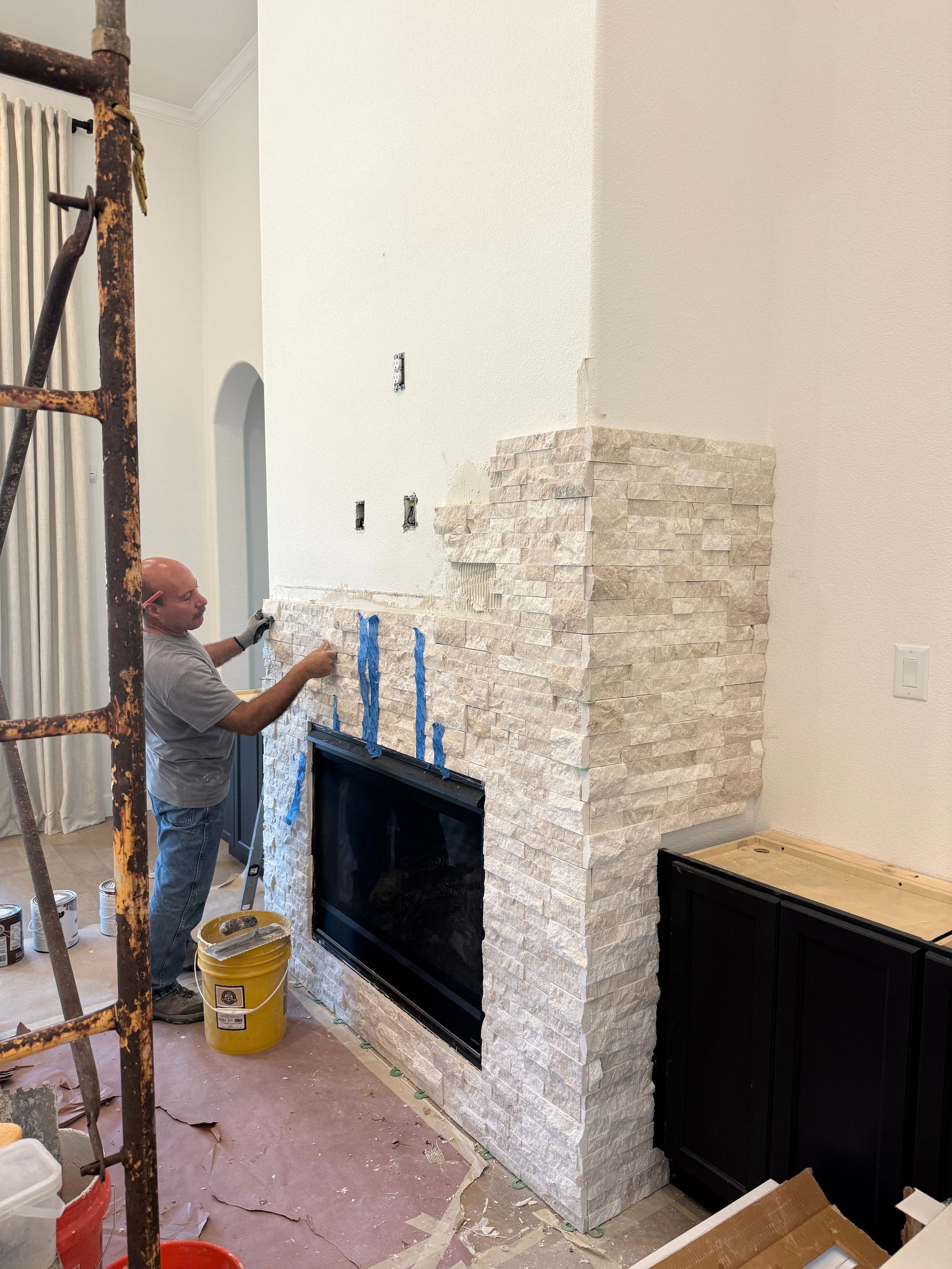 A person installs light-colored stone veneer on a fireplace surround in a room under construction.