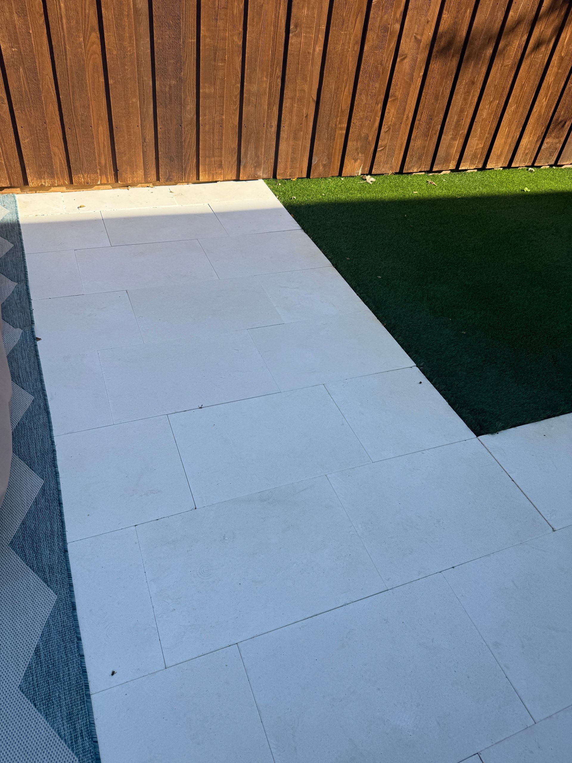 A light grey stone patio path bordered by a patterned blue rug, green artificial grass, and a wooden fence.