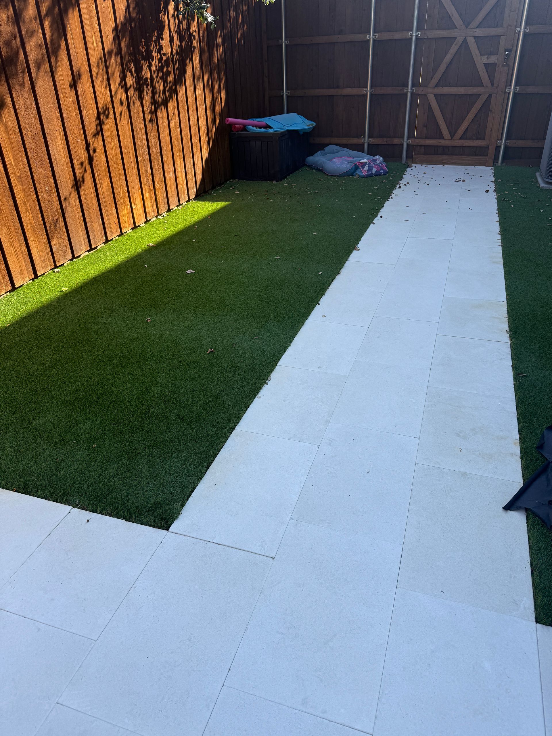 A paved pathway divides two patches of artificial grass in a backyard enclosed by a wooden fence and a gate.