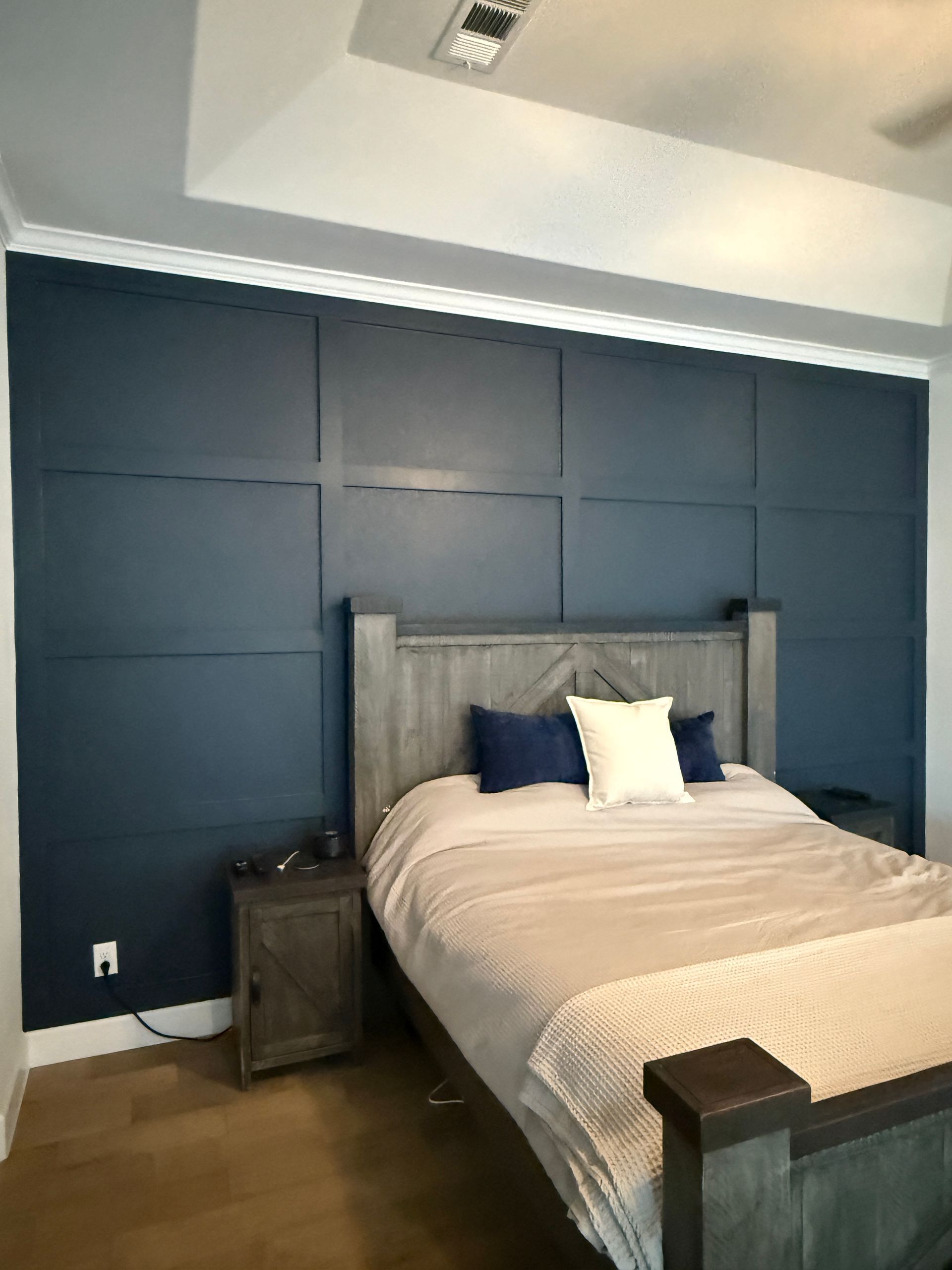 A bedroom featuring a dark blue accent wall with square molding, a wooden bed frame, and a rustic bedside table.
