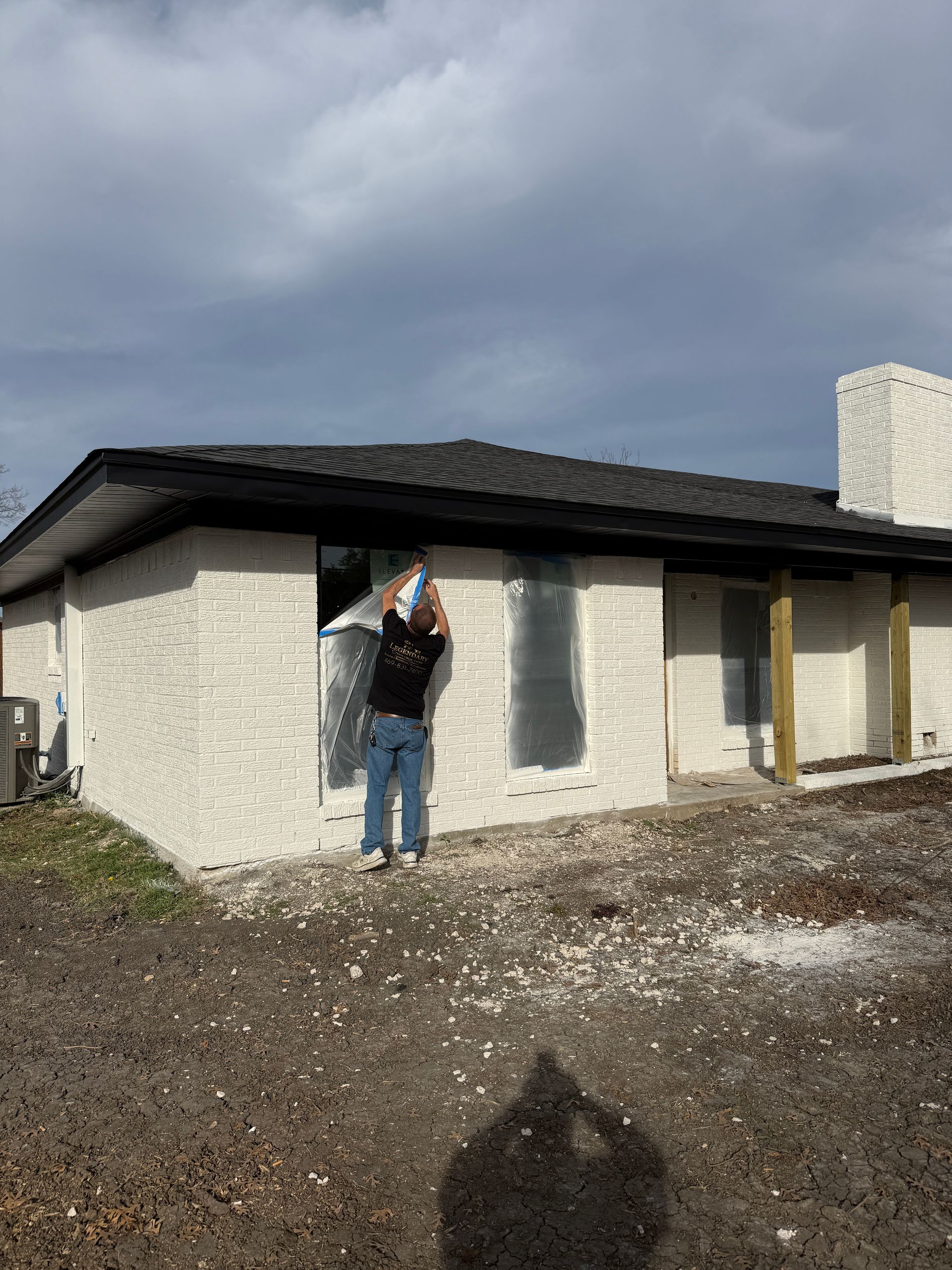 A person in a black shirt and jeans attaches plastic sheeting over a window opening on a white, stucco-finished house.