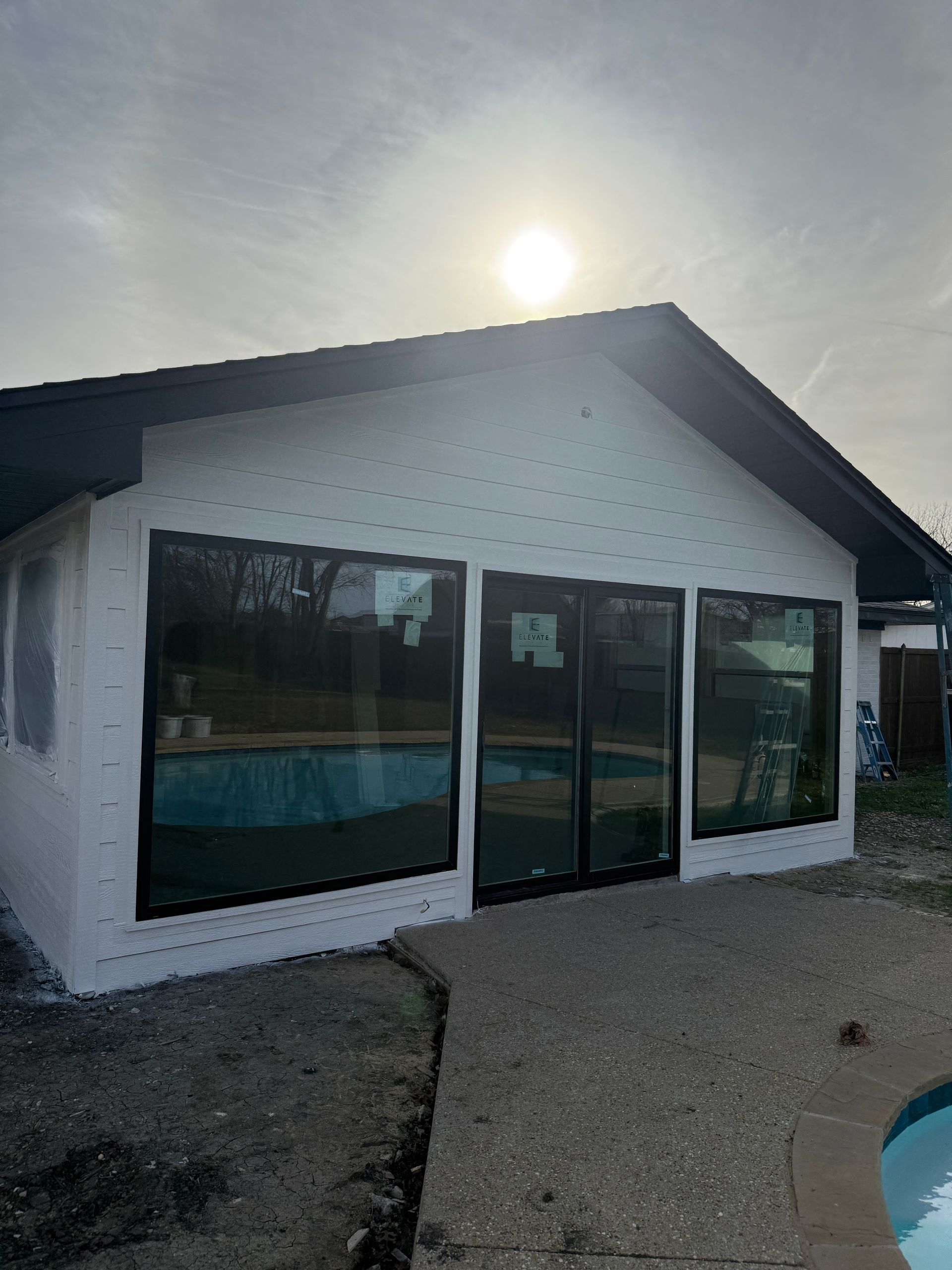 Exterior view of a house addition with a white stucco facade and new dark-framed windows overlooking a pool.