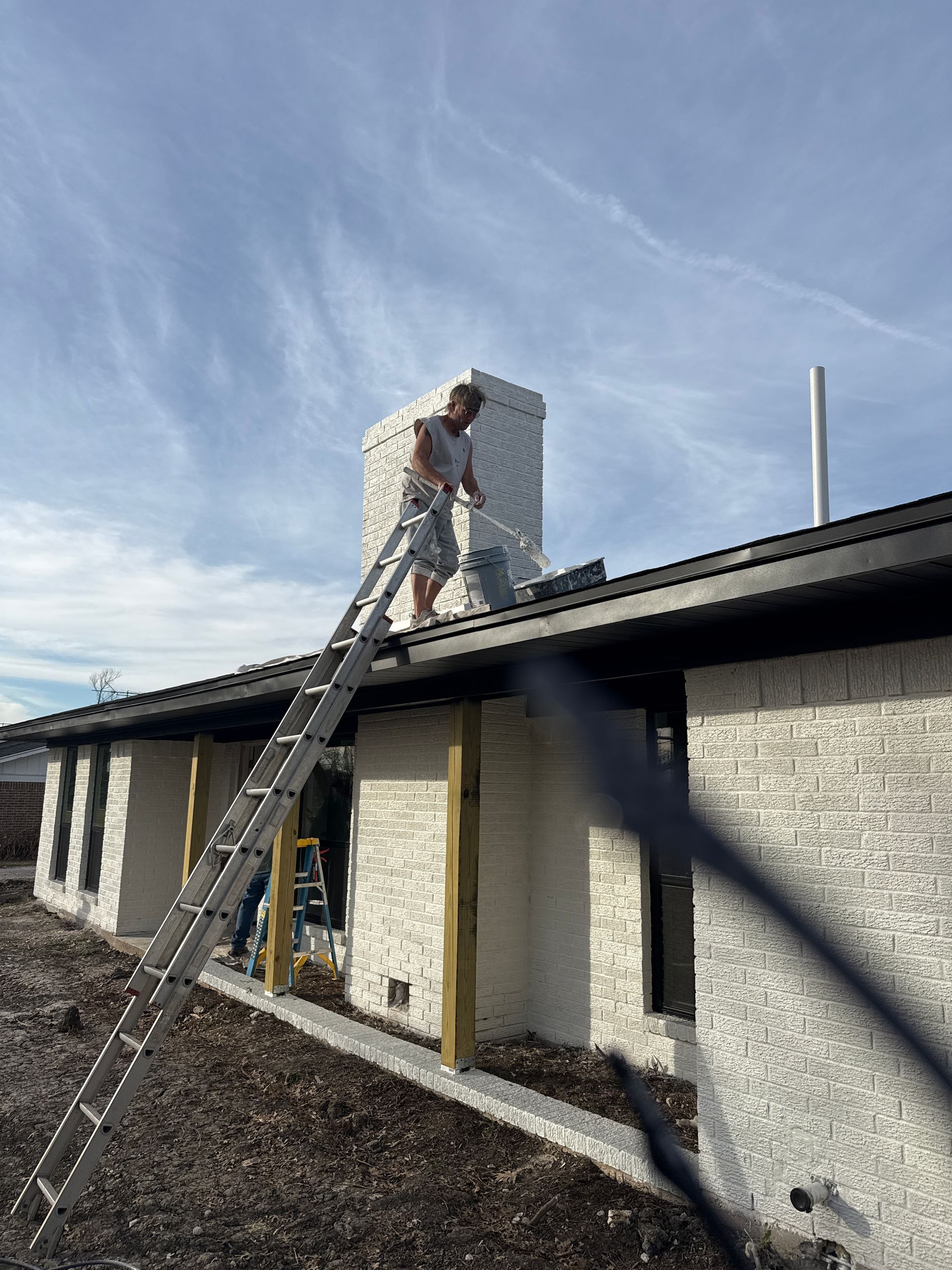 A person on a tall ladder leans against the roof edge of a white brick house, working on the chimney above.