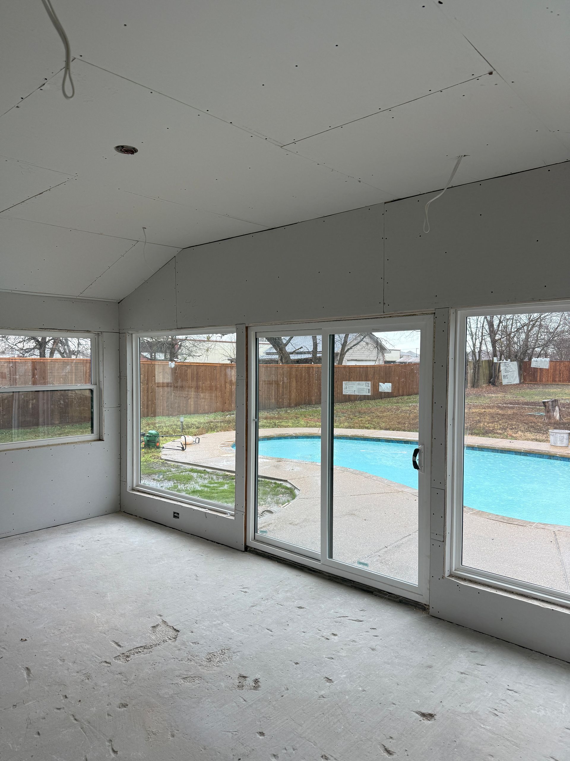A sunroom under renovation with unfinished drywall walls and ceiling, looking out onto a backyard with a swimming pool.