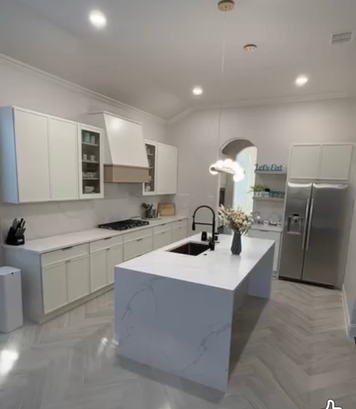 A modern, minimalist kitchen featuring white cabinets, a marble-topped island with a black sink, and herringbone flooring.