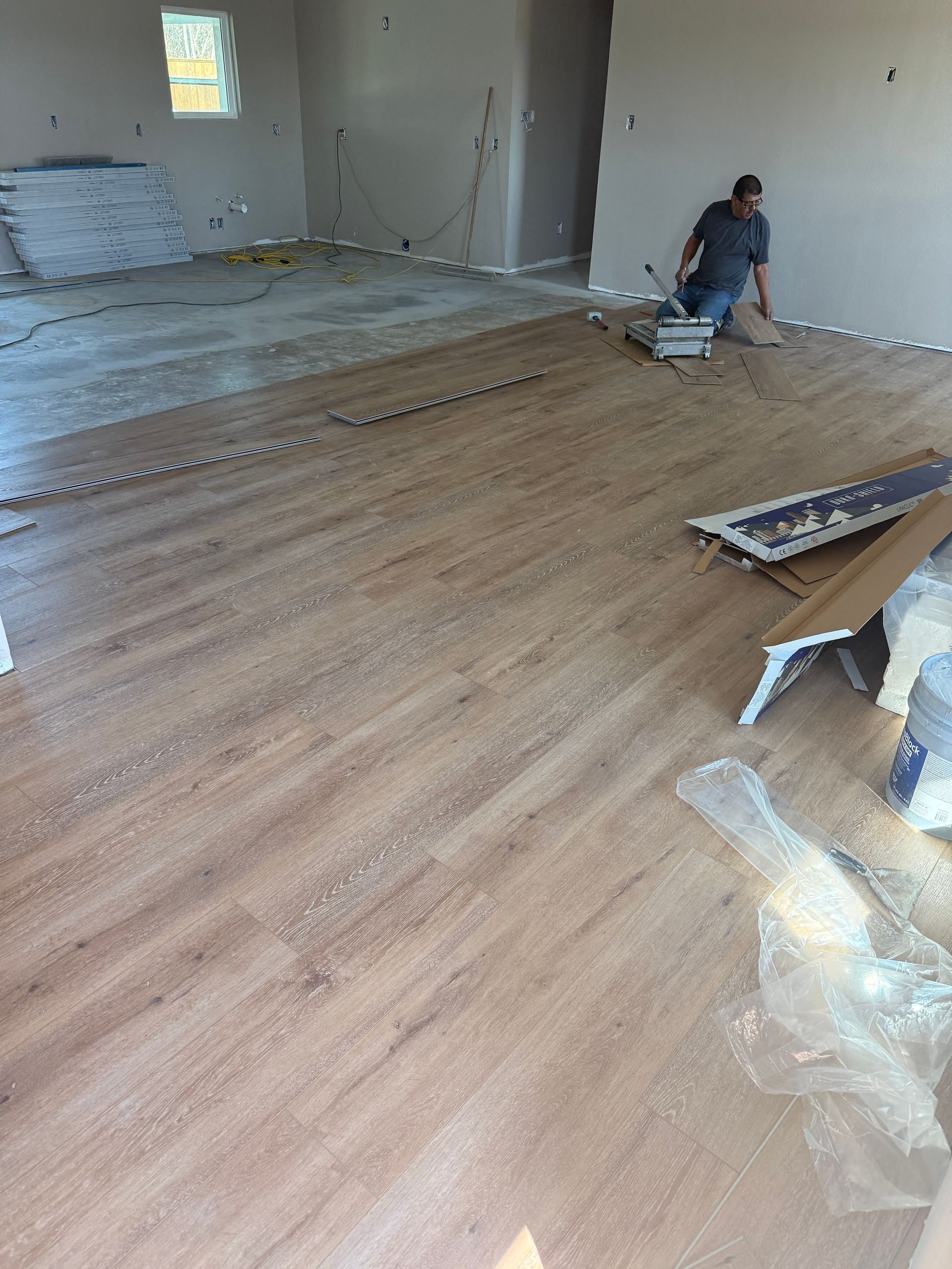 A worker installs light brown wood-look flooring in a room under construction with bags of supplies and tools nearby.