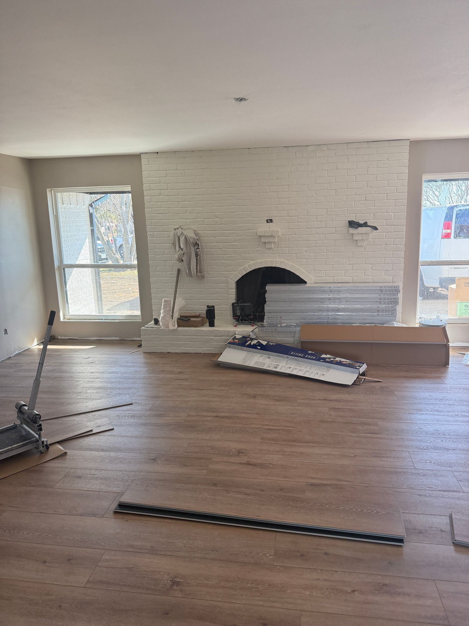 A room under renovation with new light wood-plank flooring, white brick fireplace, two windows, and construction supplies.