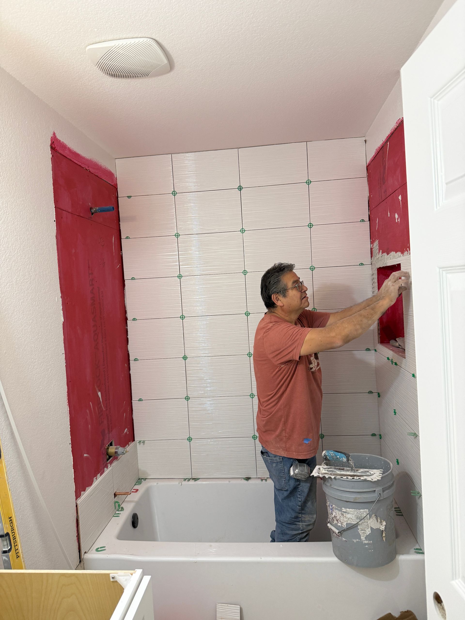 A person in a red shirt installs white, textured tiles on a shower wall prepped with red waterproof membrane.