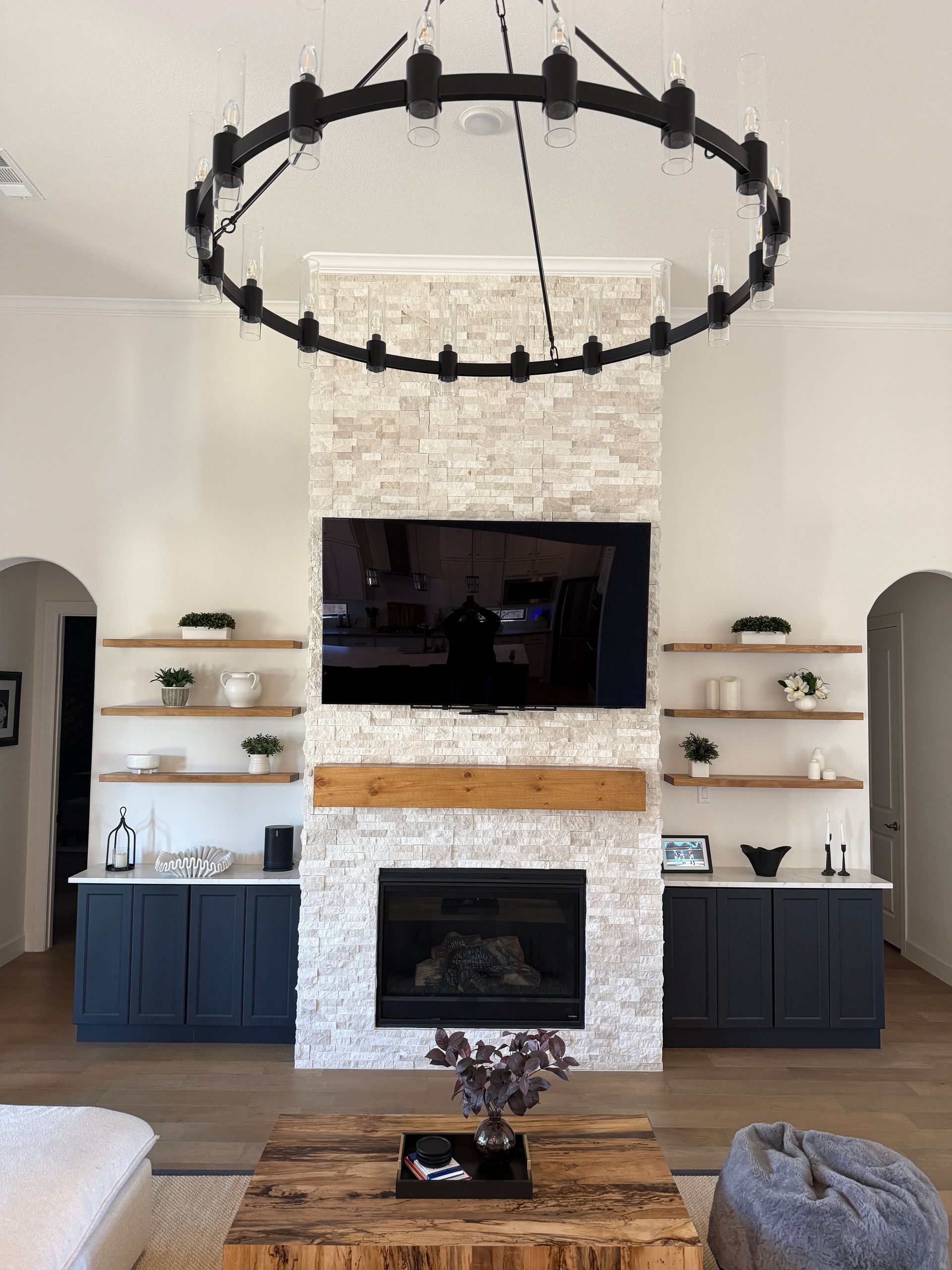 Modern living room with a stone fireplace, dark cabinets, floating shelves, and a large circular chandelier.