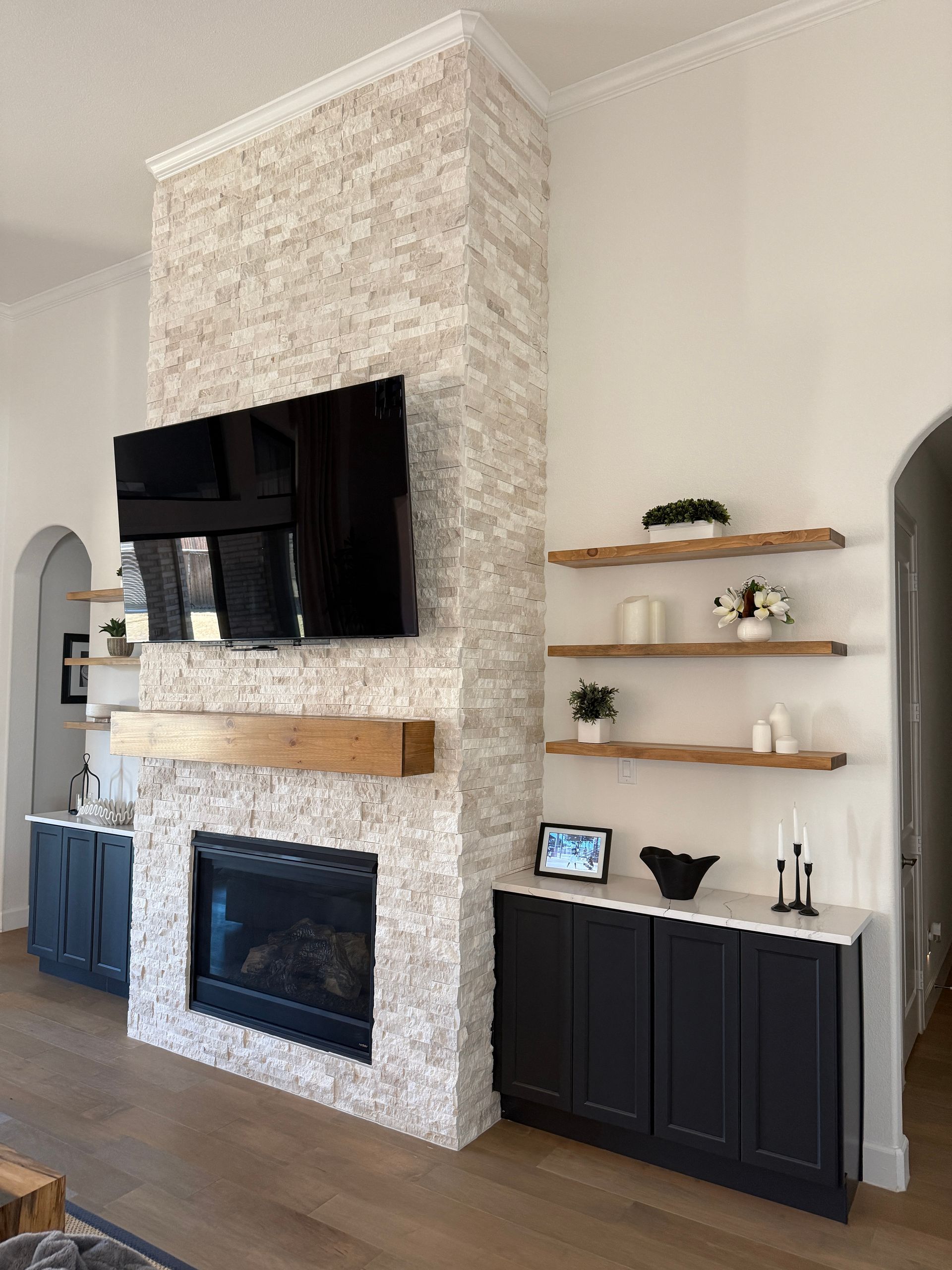 A tall stone fireplace with a television and wooden mantel, flanked by white floating shelves and dark storage cabinets.