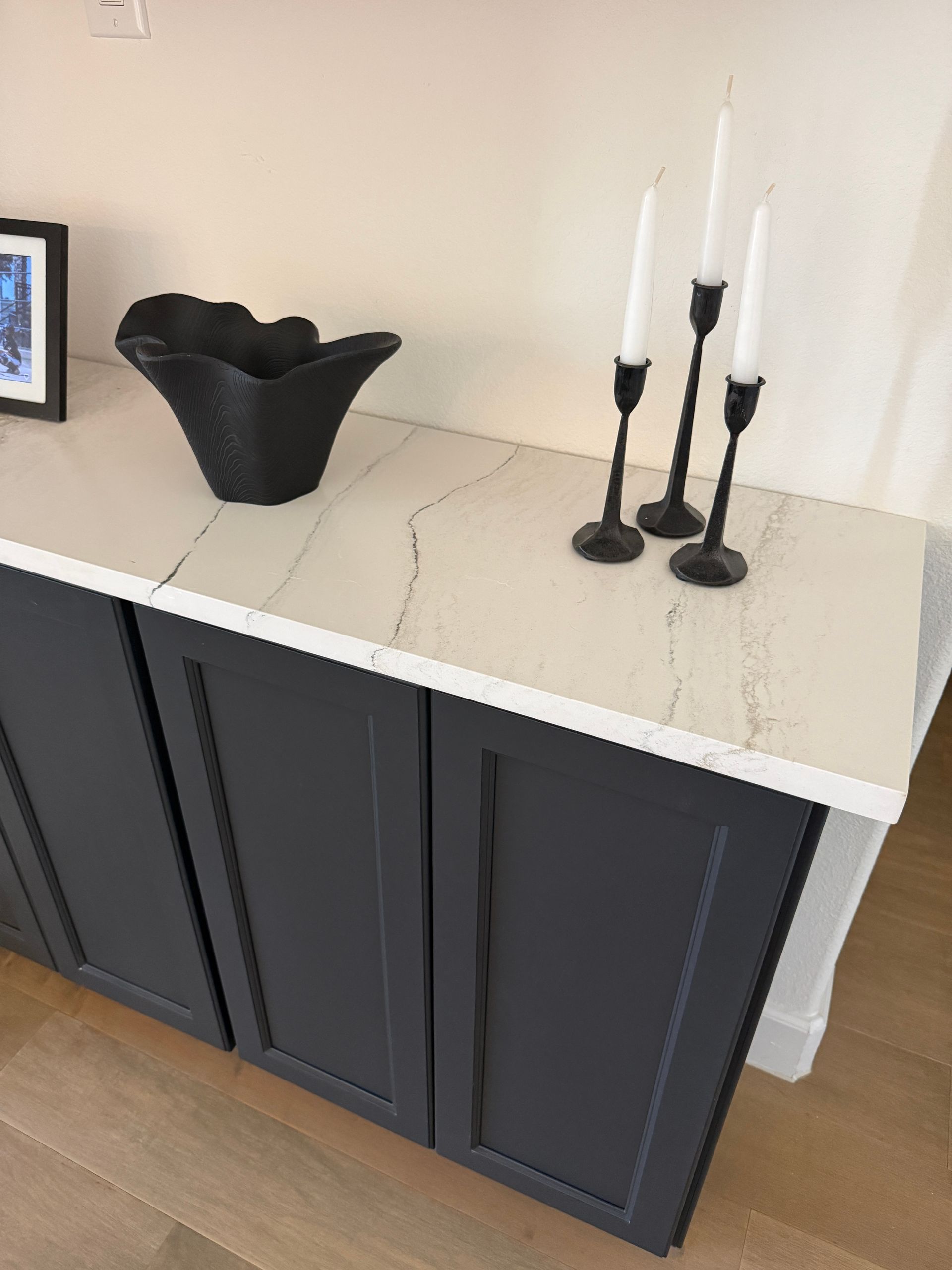 A stone-topped cabinet with dark blue cupboards, a black abstract bowl, and three black candle holders with white candles.